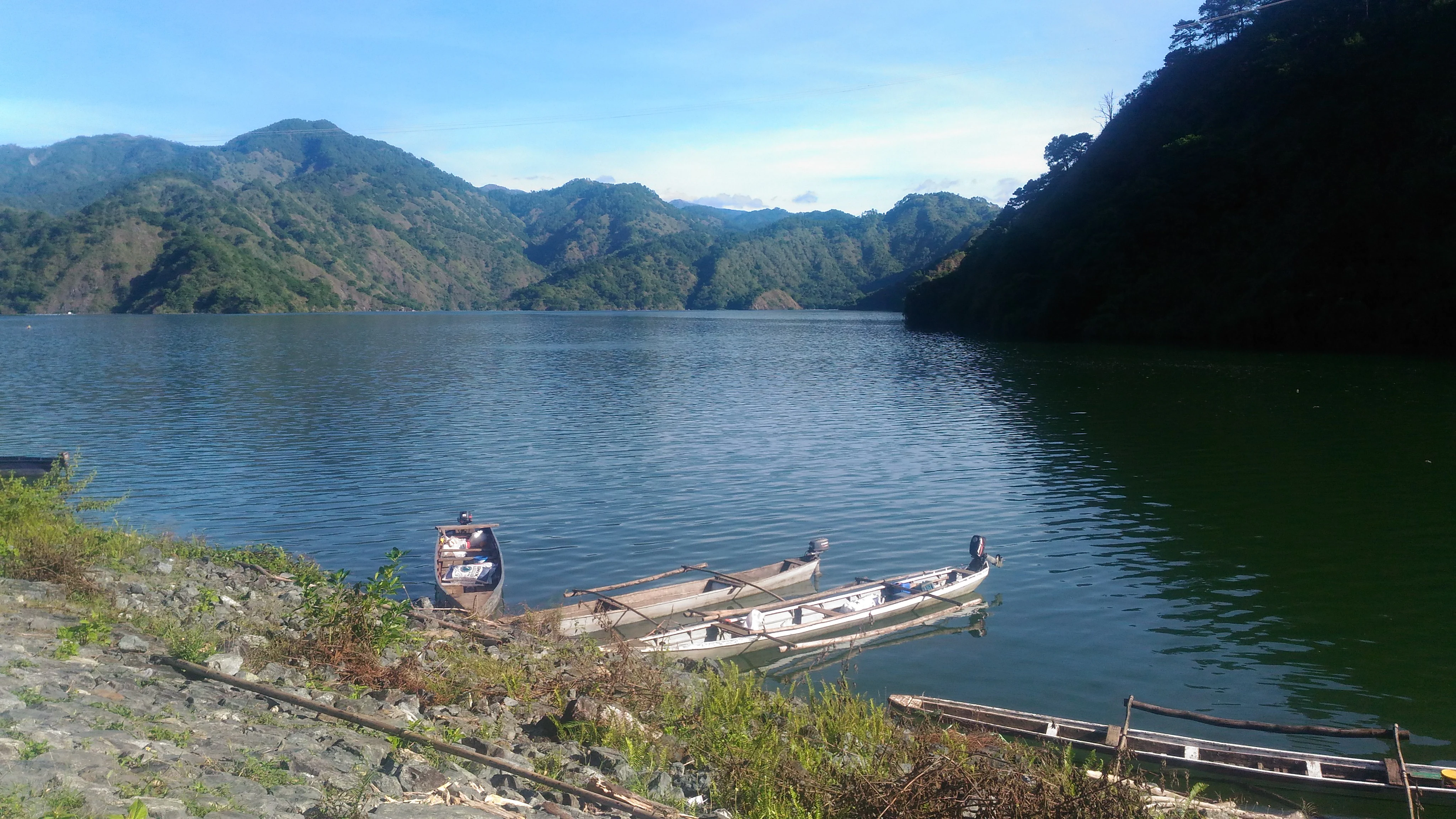 A calm, wide lake surrounded by steep, tree-covered mountains, with several small traditional boats docked along the rocky foreground shore.