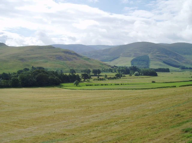 A wide view of a pastoral valley featuring rolling green hills and patches of trees, with golden fields in the foreground and mountains in the background under a cloudy sky.