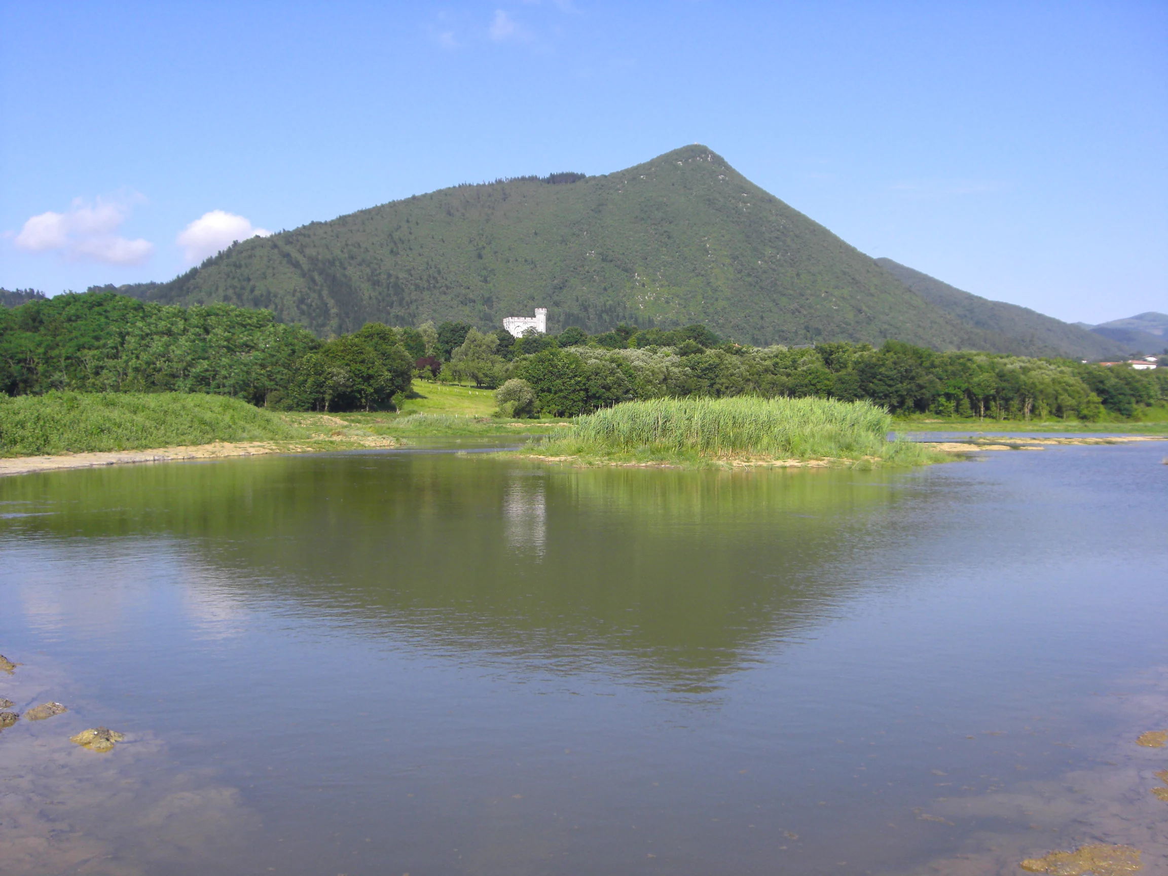 A calm, still lagoon reflecting a nearby grassy hill and a small white tower structure nestled among trees in the background under a blue sky.