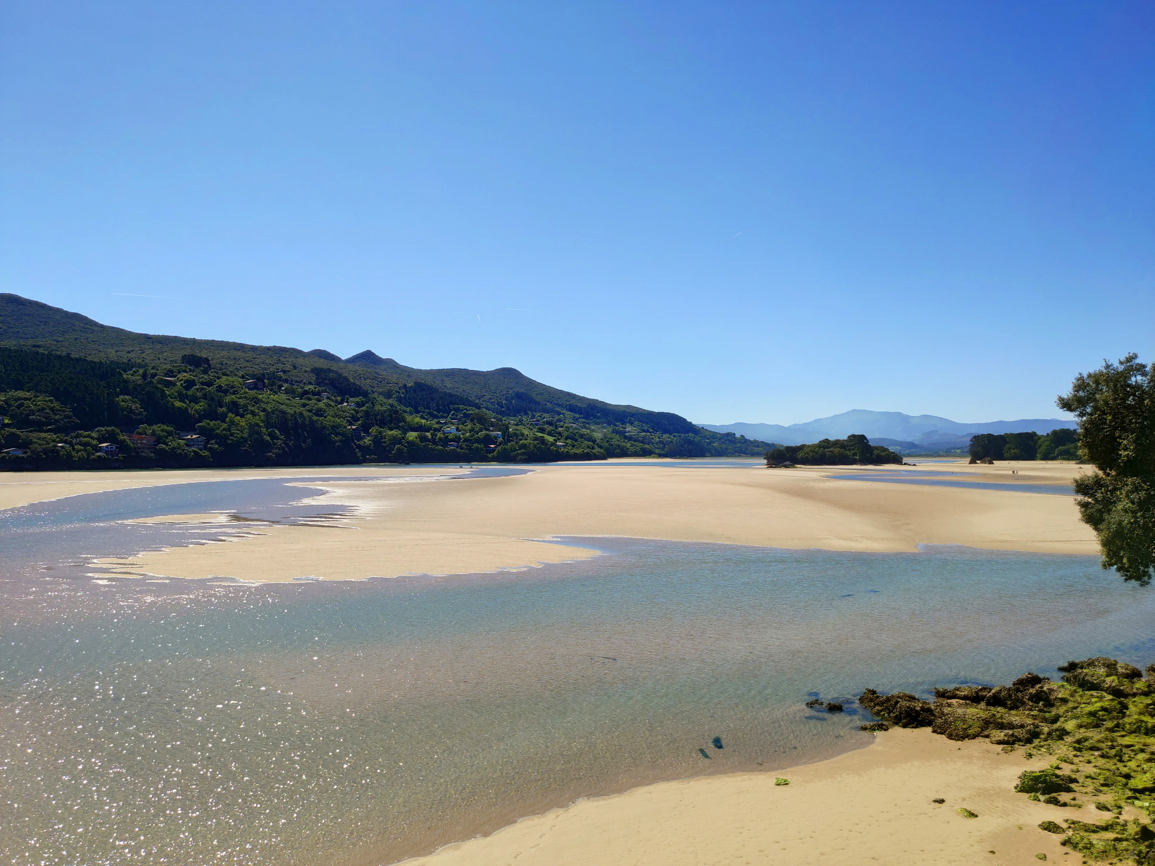 A wide view of a large sandy tidal estuary at low tide, with low green hills in the background under a clear blue sky.