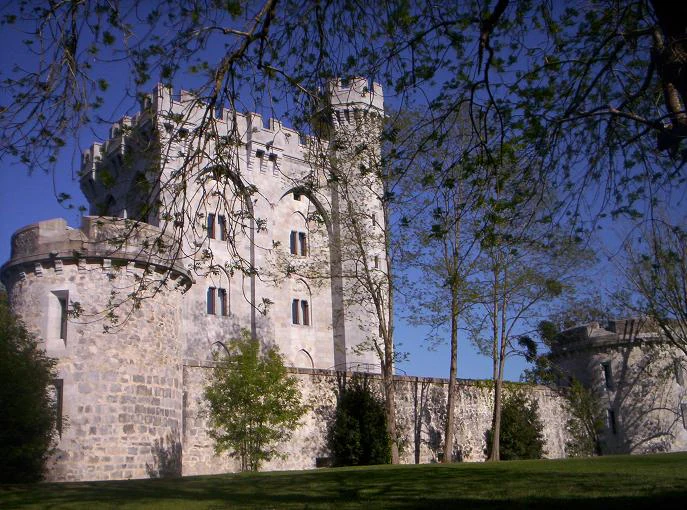 A medieval-style stone castle or tower with crenellated walls and a prominent tower, surrounded by trees and a grassy foreground.