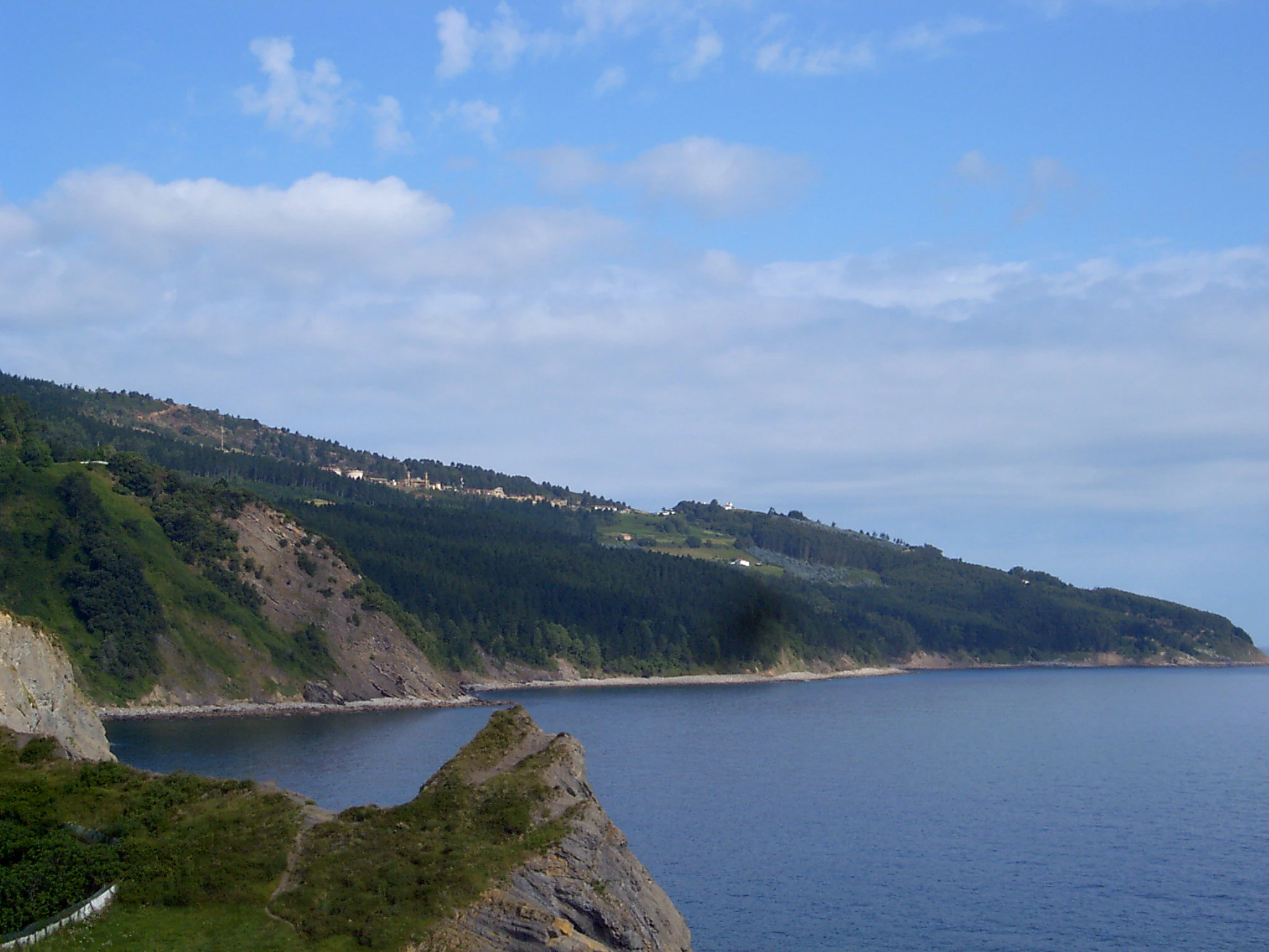A wide shot of the rocky, green coastline at Matxitxako Cape meeting the calm deep blue ocean under a partly cloudy sky.