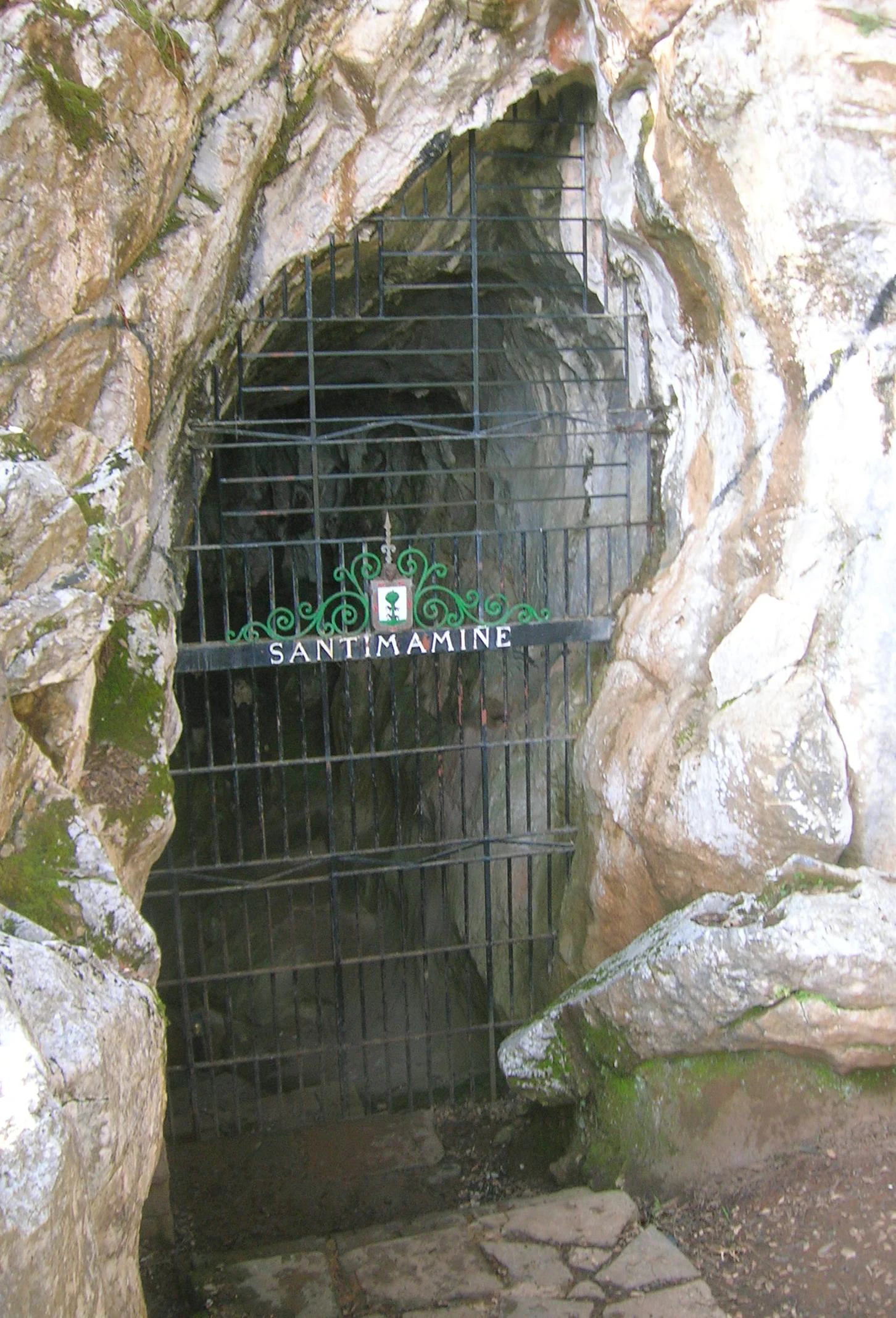 A gated entrance to a natural limestone cave opening in a cliff face; a sign on the metal gate reads SANTIMAMINE.