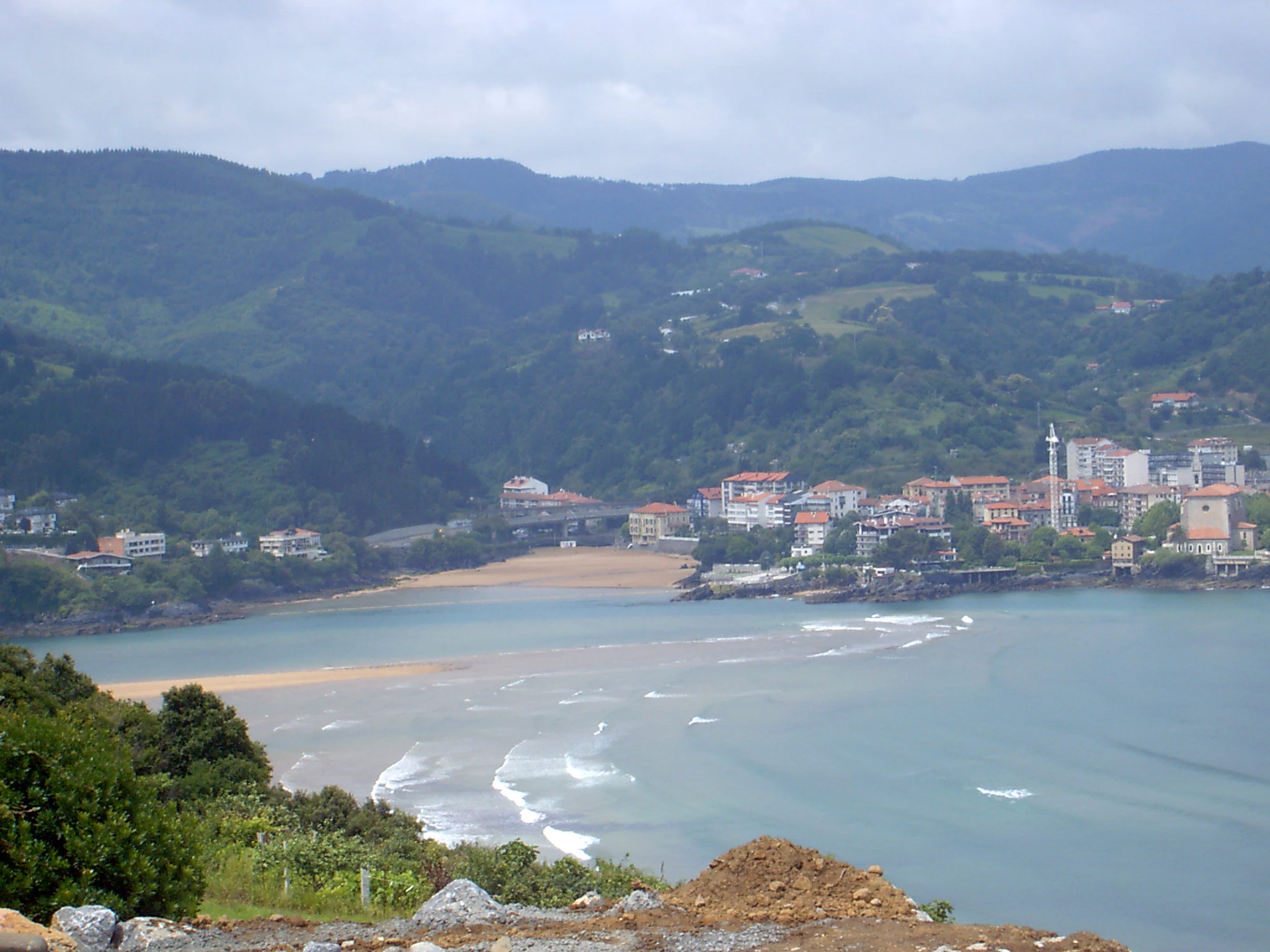 A coastal landscape shot looking across the Mundaka estuary with the town of Mundaka visible on the shore and lush green hills in the background.