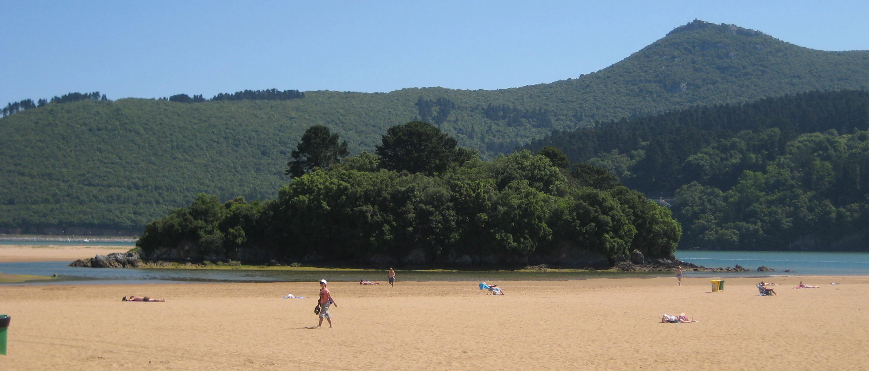 A sandy beach in the foreground leads to a small, tree-covered island in the water, with rolling green hills and a mountain peak in the background.