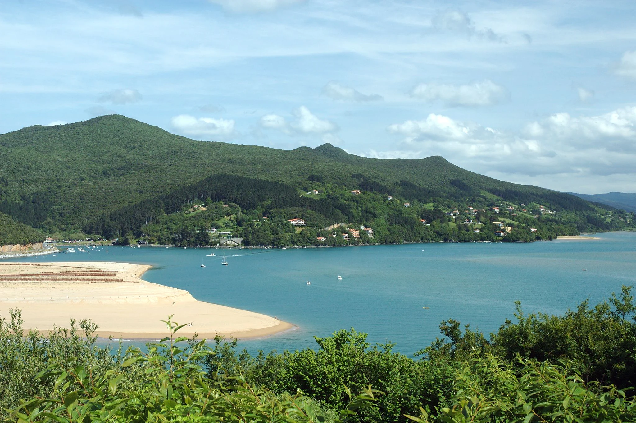 A wide shot showing a sandy beach curving into a wide blue estuary, backed by green hills and forests under a partly cloudy sky.