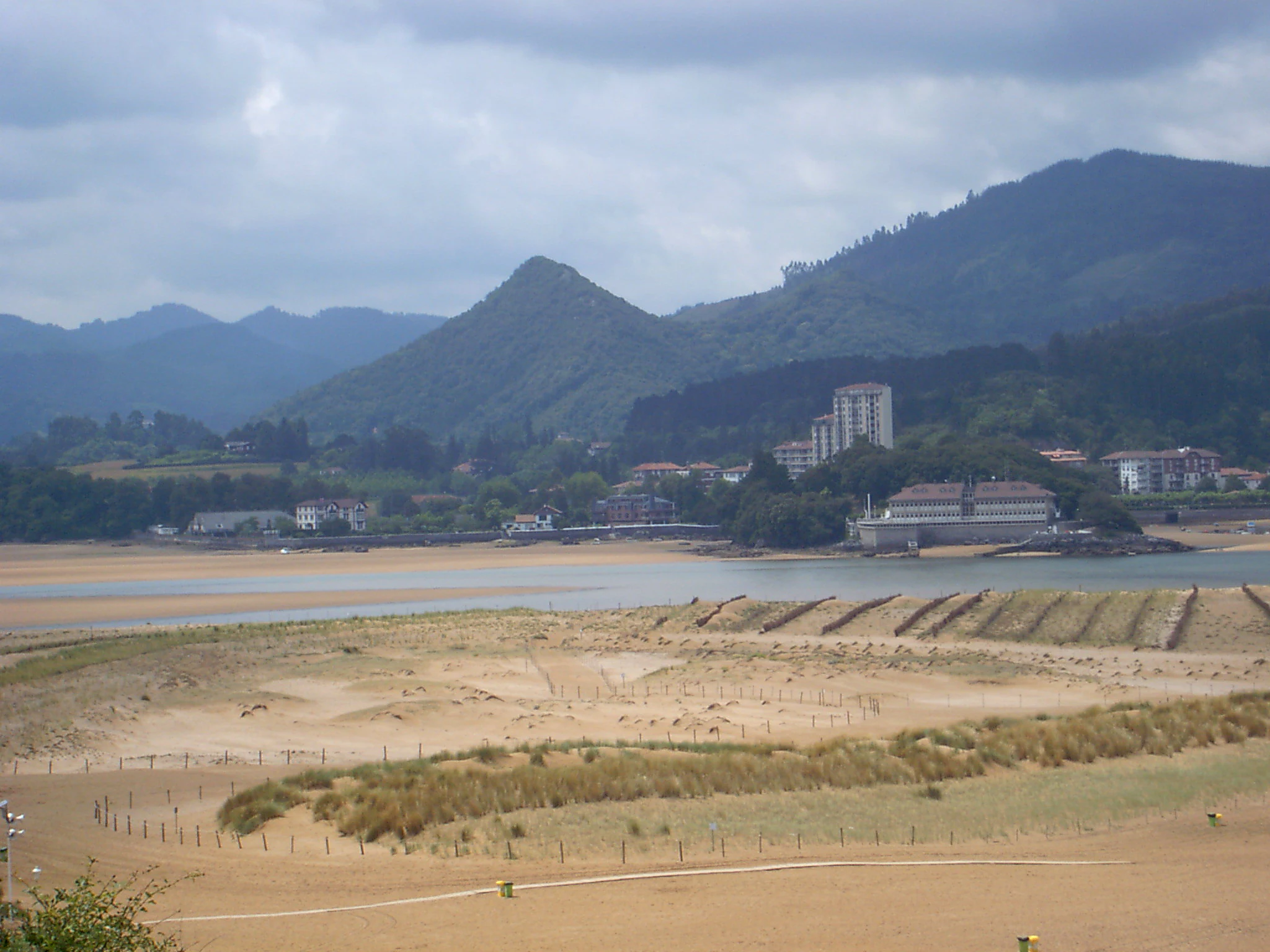 A wide shot of a sandy beach in the foreground leading to the Oka river estuary, with hills and a small town in the background under a cloudy sky.