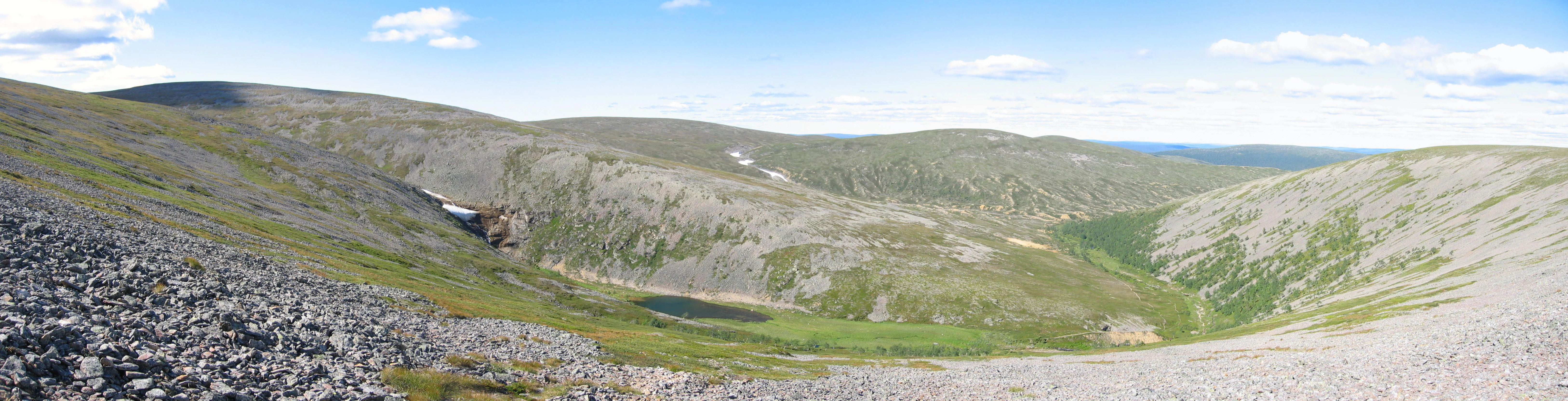 Wide panoramic view of a valley with rocky hillsides, a small lake, and green terrain under a partly cloudy sky