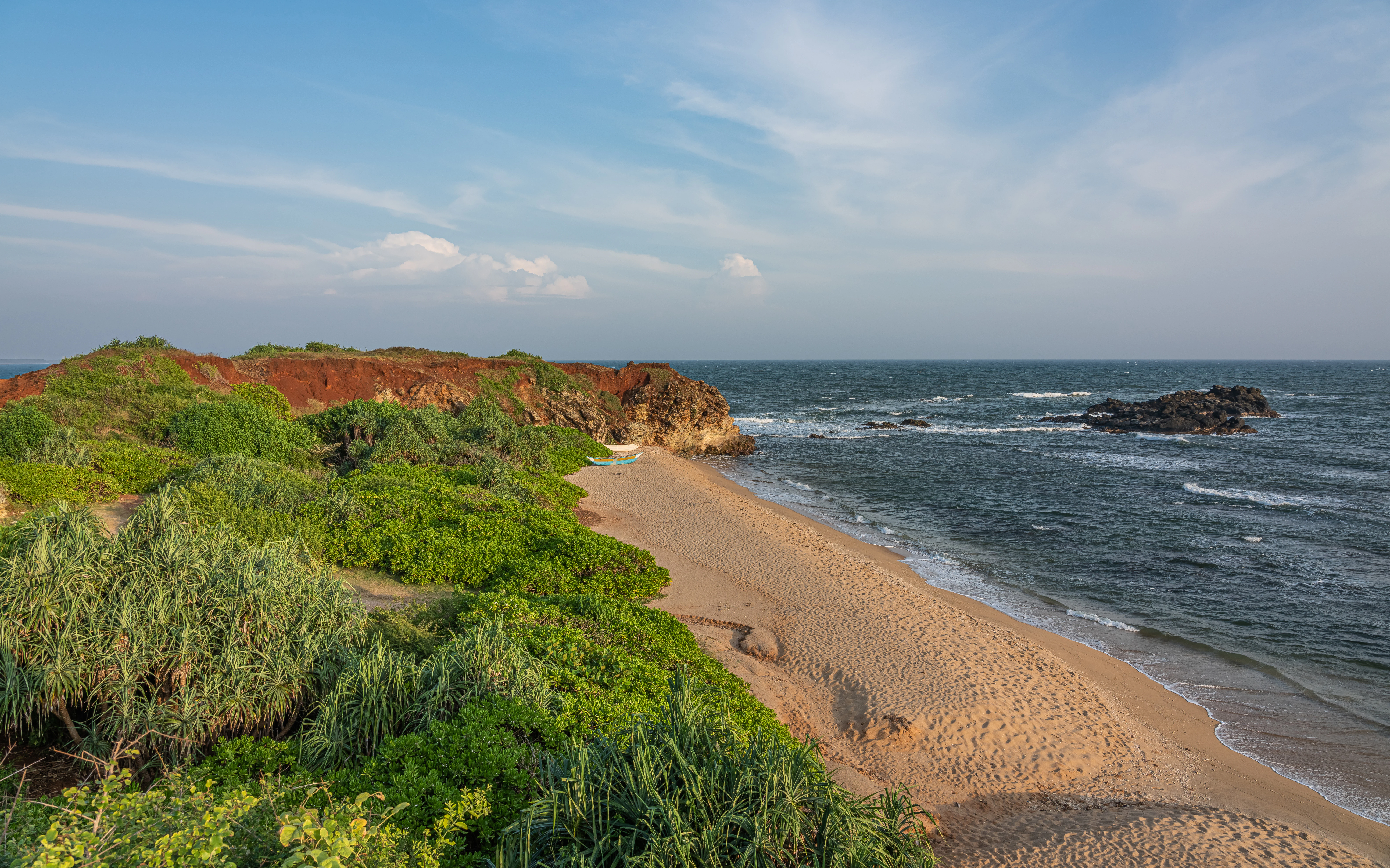 Sandy beach with waves, rocky cliffs covered in vegetation, and ocean under a partly cloudy sky.