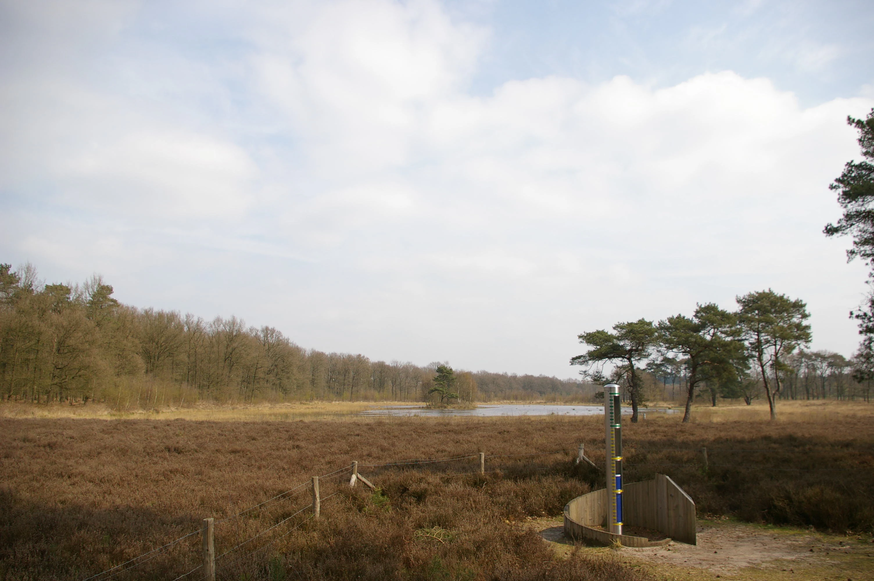 Wide landscape view of Utrechtse Heuvelrug National Park featuring brown heathland in the foreground, a body of water (Egelmeer) in the middle ground, scattered trees, and a partly cloudy sky