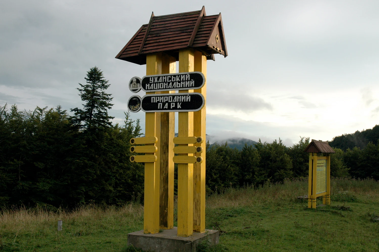 Yellow signpost with two black directional signs reading 'Ужанський національний природний парк' and 'Природний парк' under a small roof, set in a grassy field with forest and hills in the background