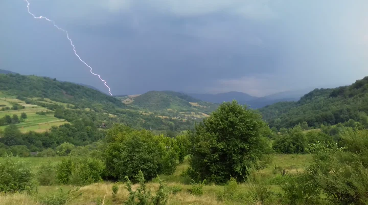 A lightning bolt strikes over a mountainous landscape with green forests and rolling hills under a dark stormy sky