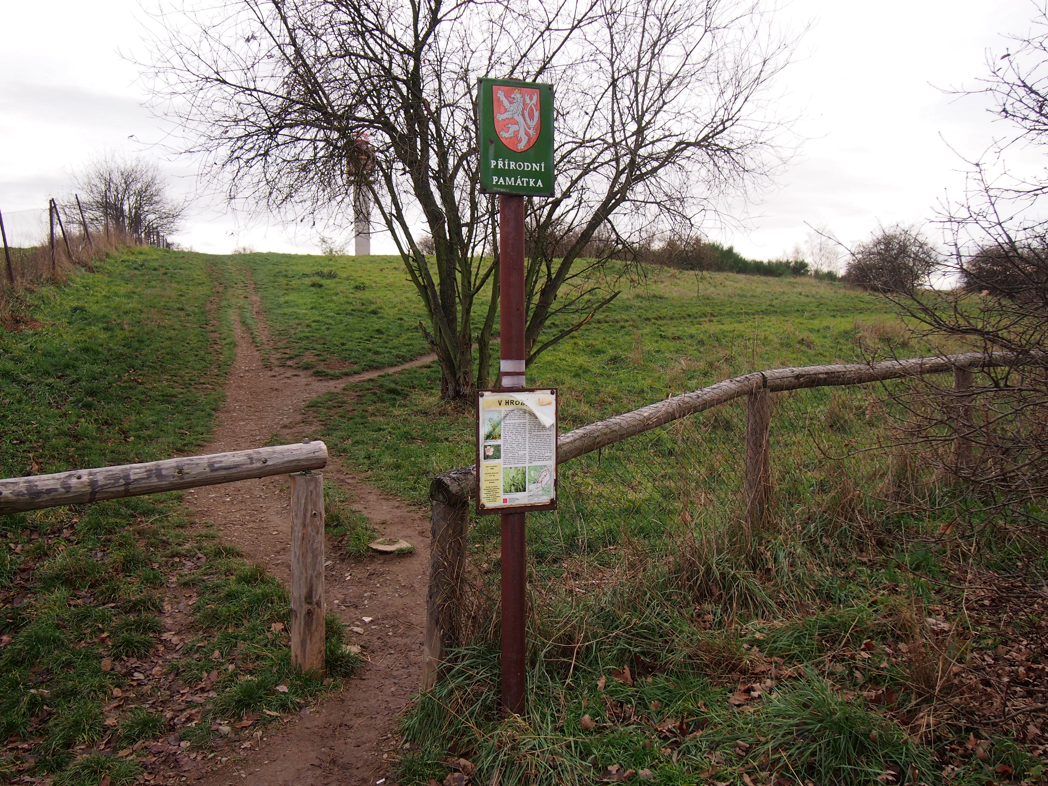 A signpost for the V hrobech natural monument in Czechia, featuring a national emblem at the top and an information board below, situated at the entrance of a grassy trail with wooden fencing.