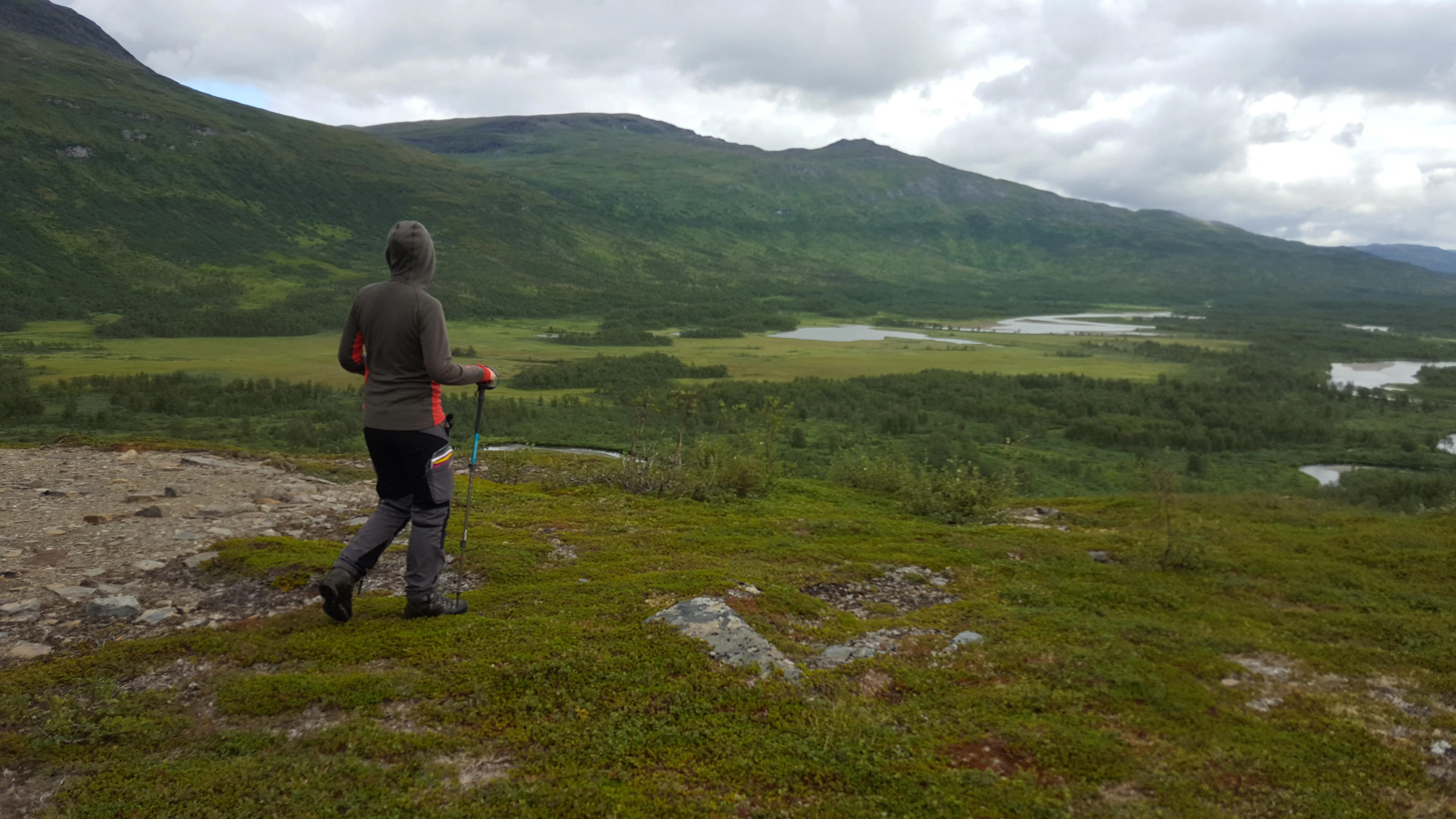 Person hiking with walking stick on grassy hillside overlooking valley with water bodies and mountains in background