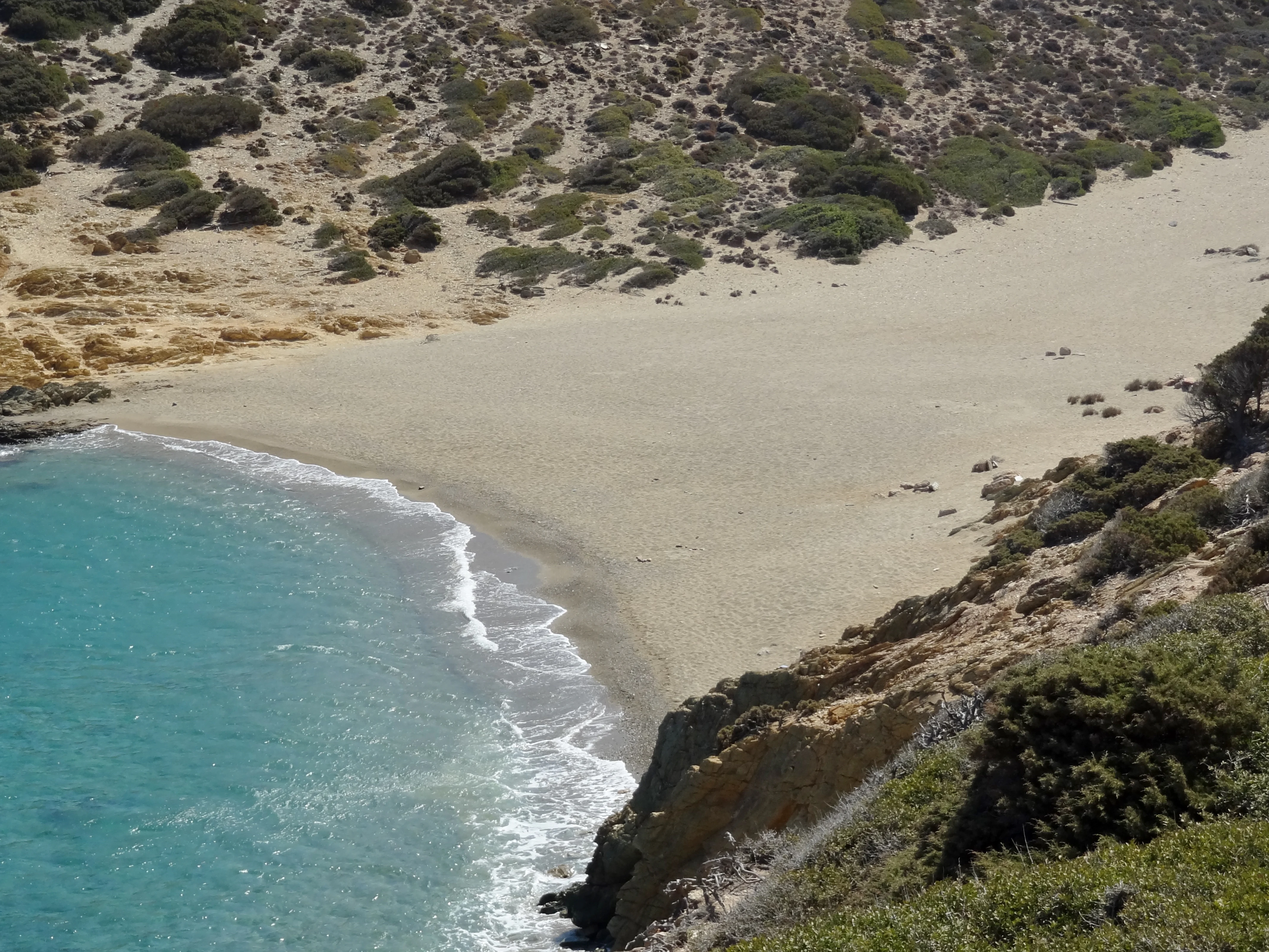 A high-angle view of a small, sandy beach tucked into a cove with turquoise water and rocky, scrub-covered hillsides.