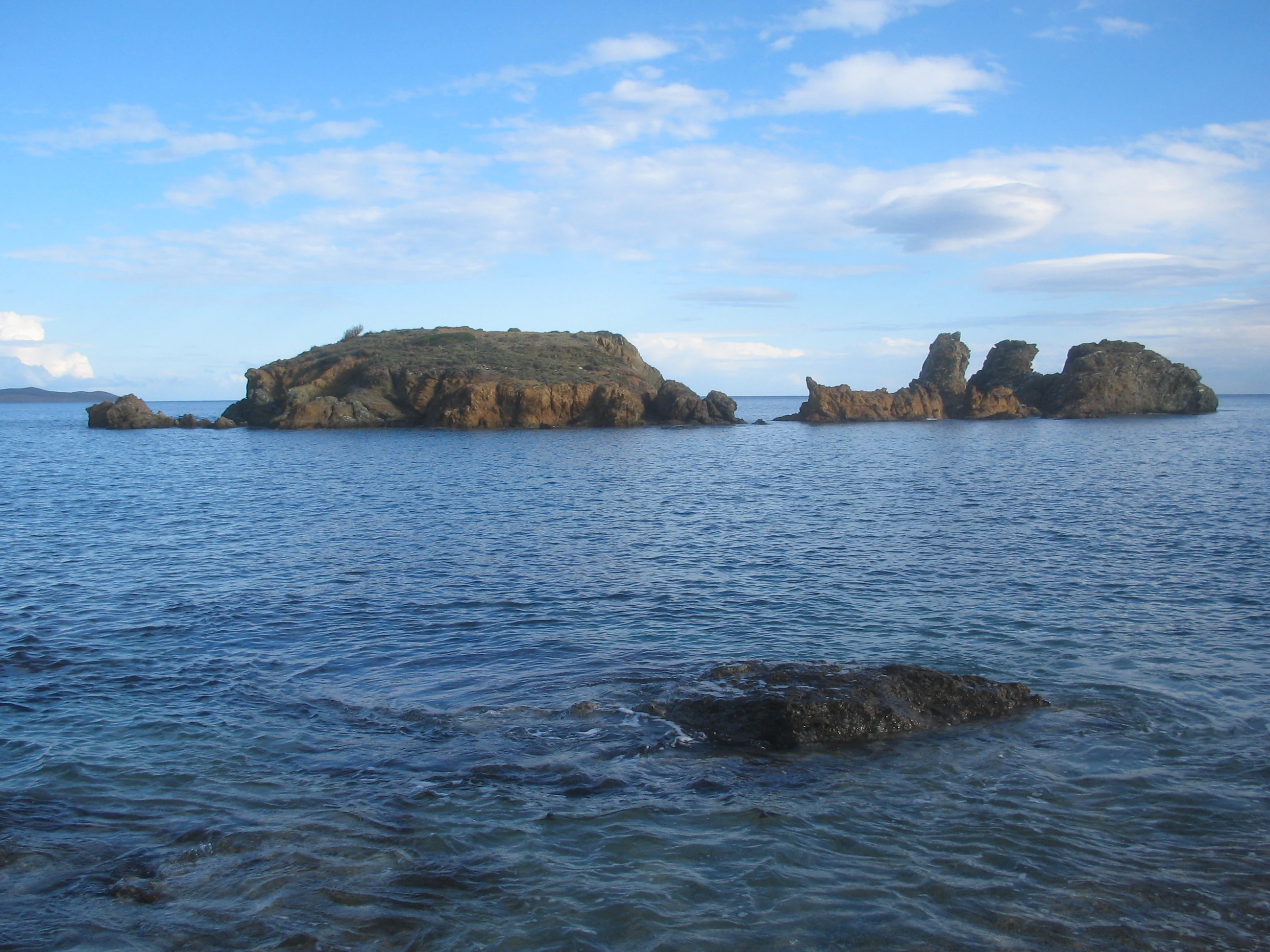 A view across calm blue coastal waters toward a small, elongated rocky island and smaller formations, under a partly cloudy sky.