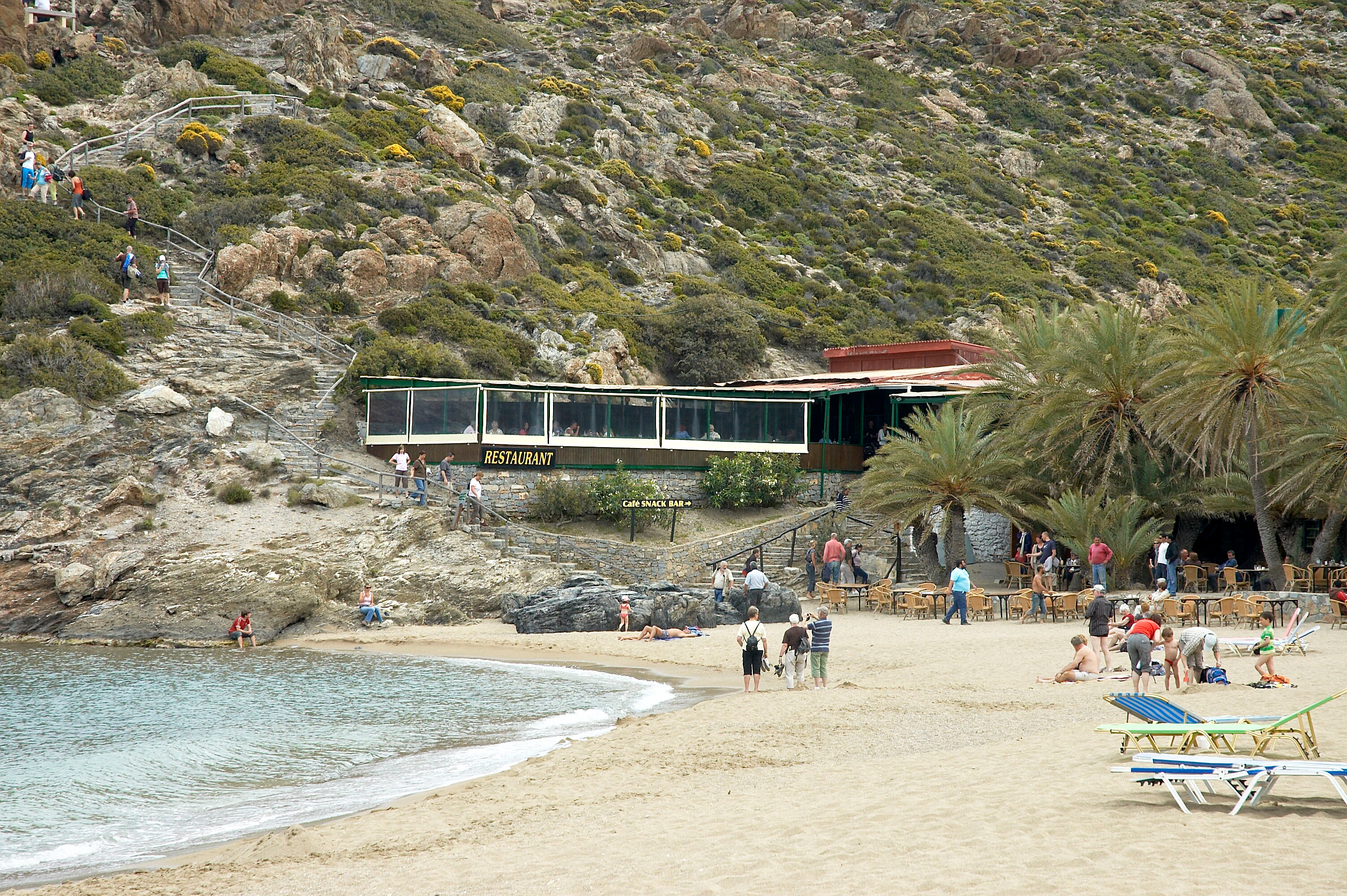 A wide view of a sandy beach with people, a restaurant building, and palm trees at the base of a rocky hillside, with a staircase leading up the slope.