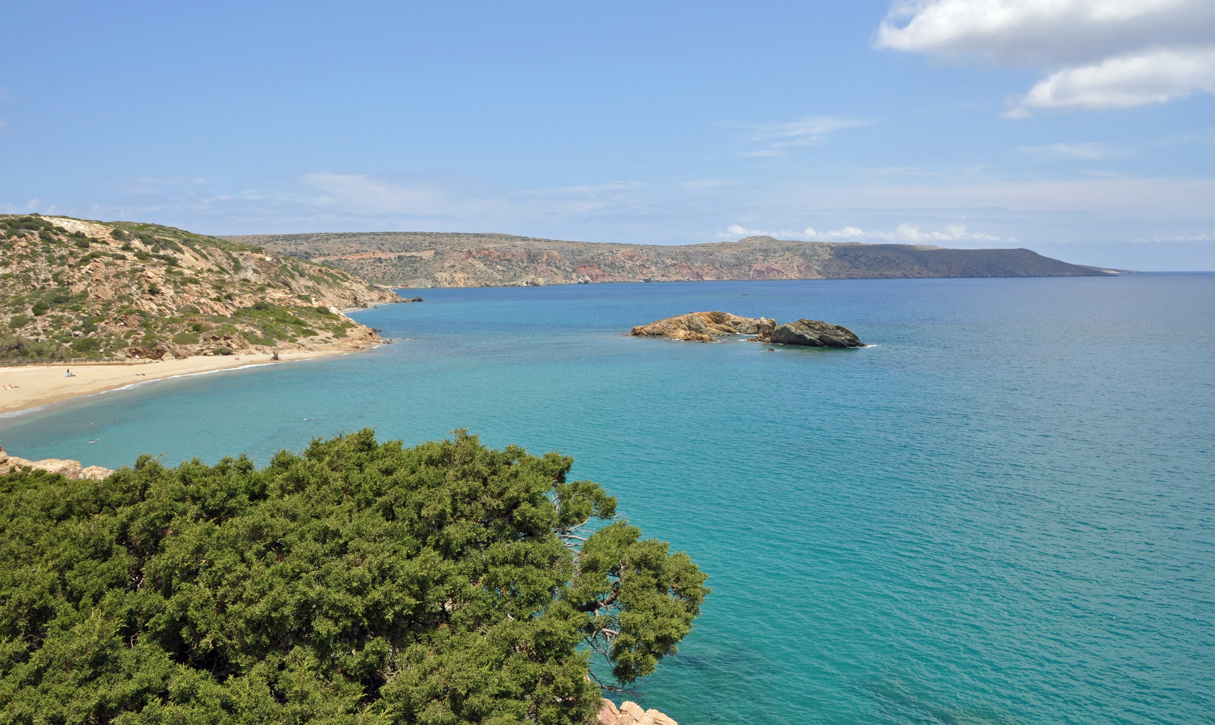 A wide angle view of the sandy beach and turquoise waters of Vai Bay, with dense green palm vegetation in the foreground and rocky hills in the background.