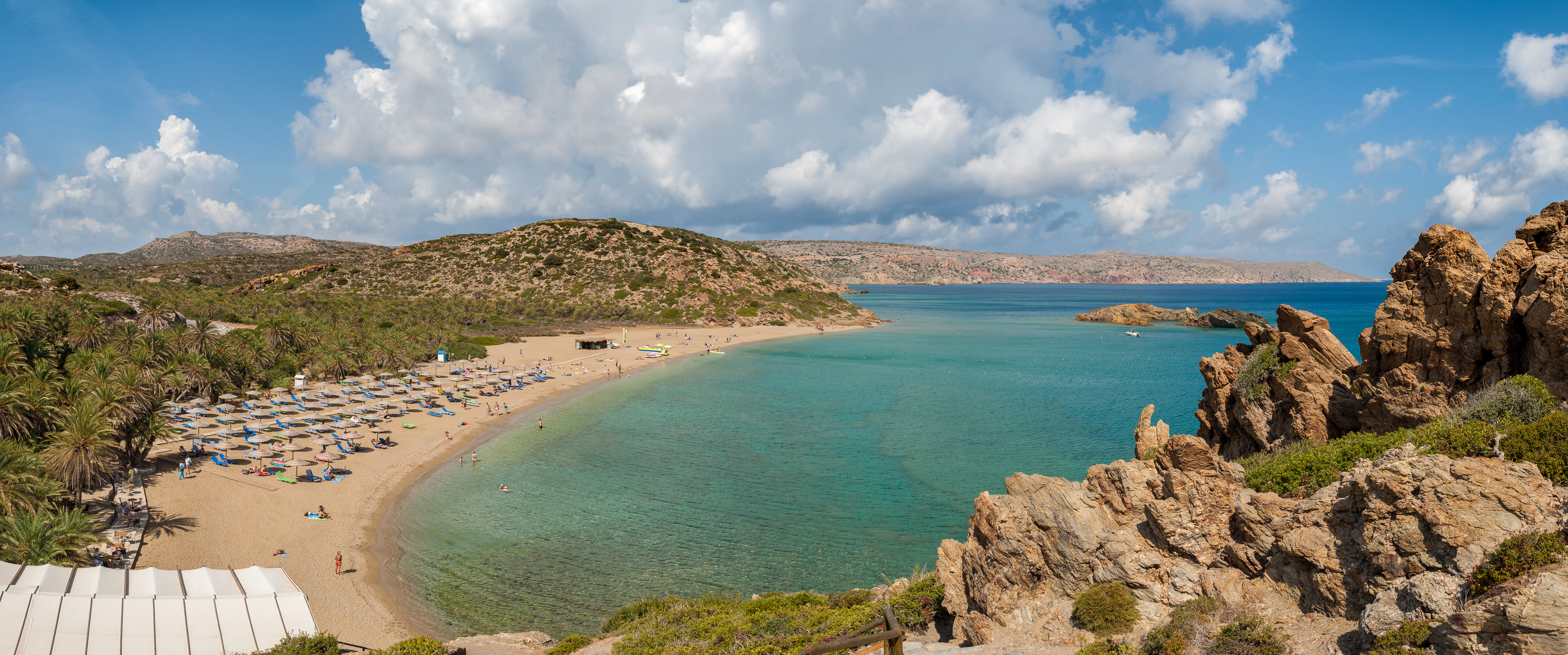 Panoramic view of a sandy beach lined with palm trees meeting the sea, with rugged rocky cliffs in the foreground and hills in the background under a blue sky with scattered clouds.