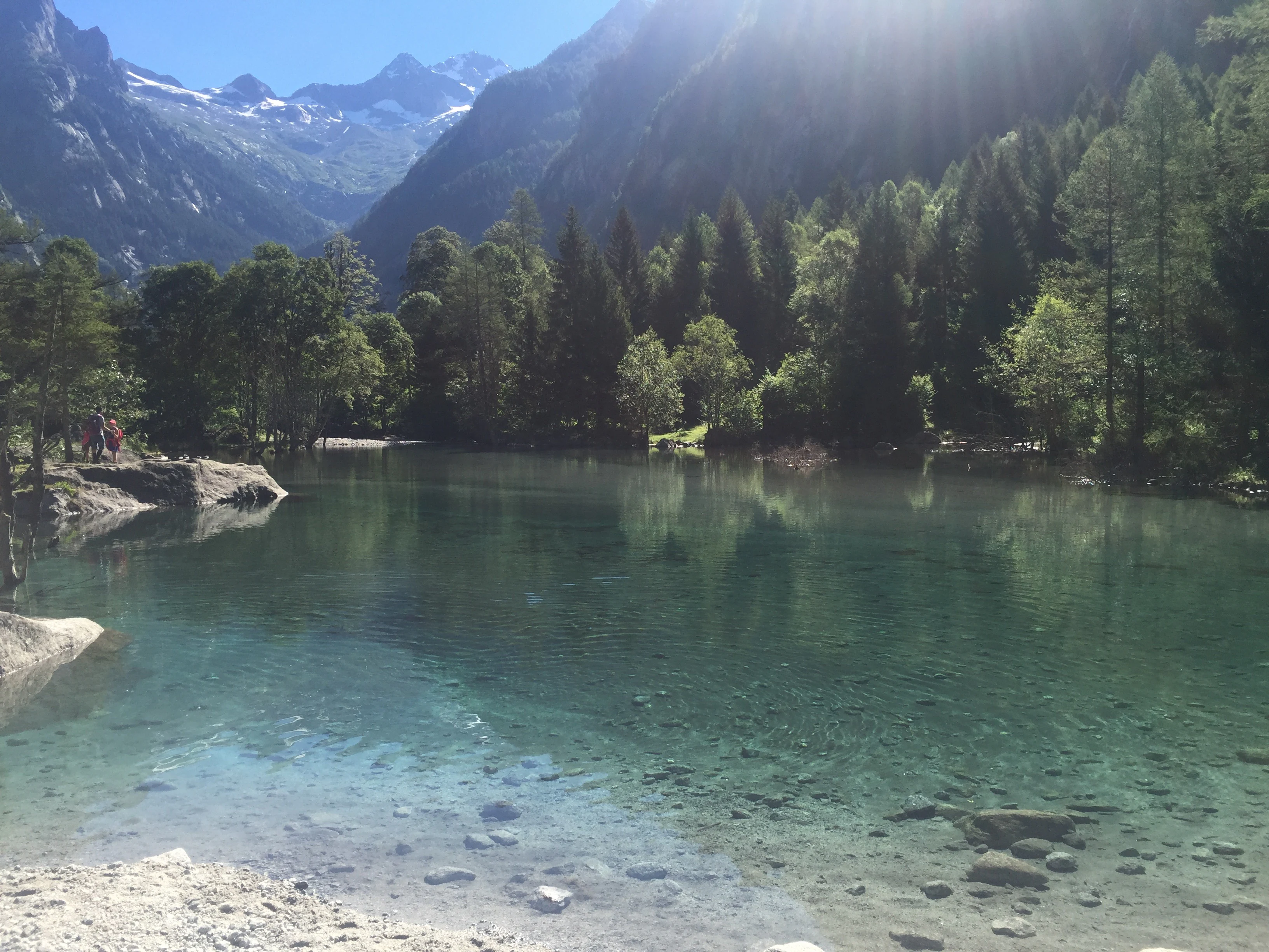 A bright, clear alpine lake with turquoise water reflecting surrounding pine trees, set against a backdrop of steep, rugged mountains under a sunny sky.