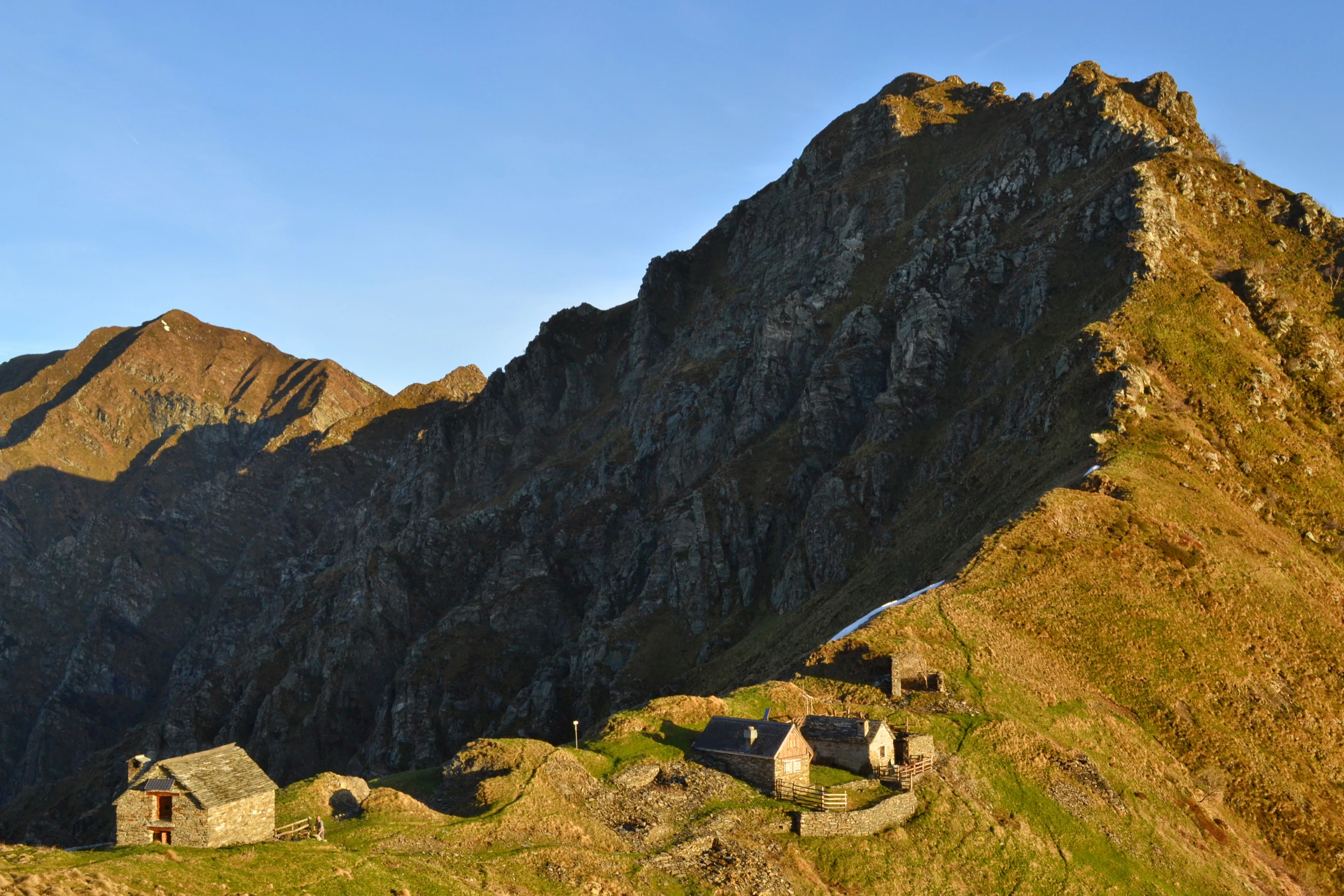 Stone bivouac hut with sloped roof on grassy mountain slope, surrounded by rocky peaks under clear blue sky