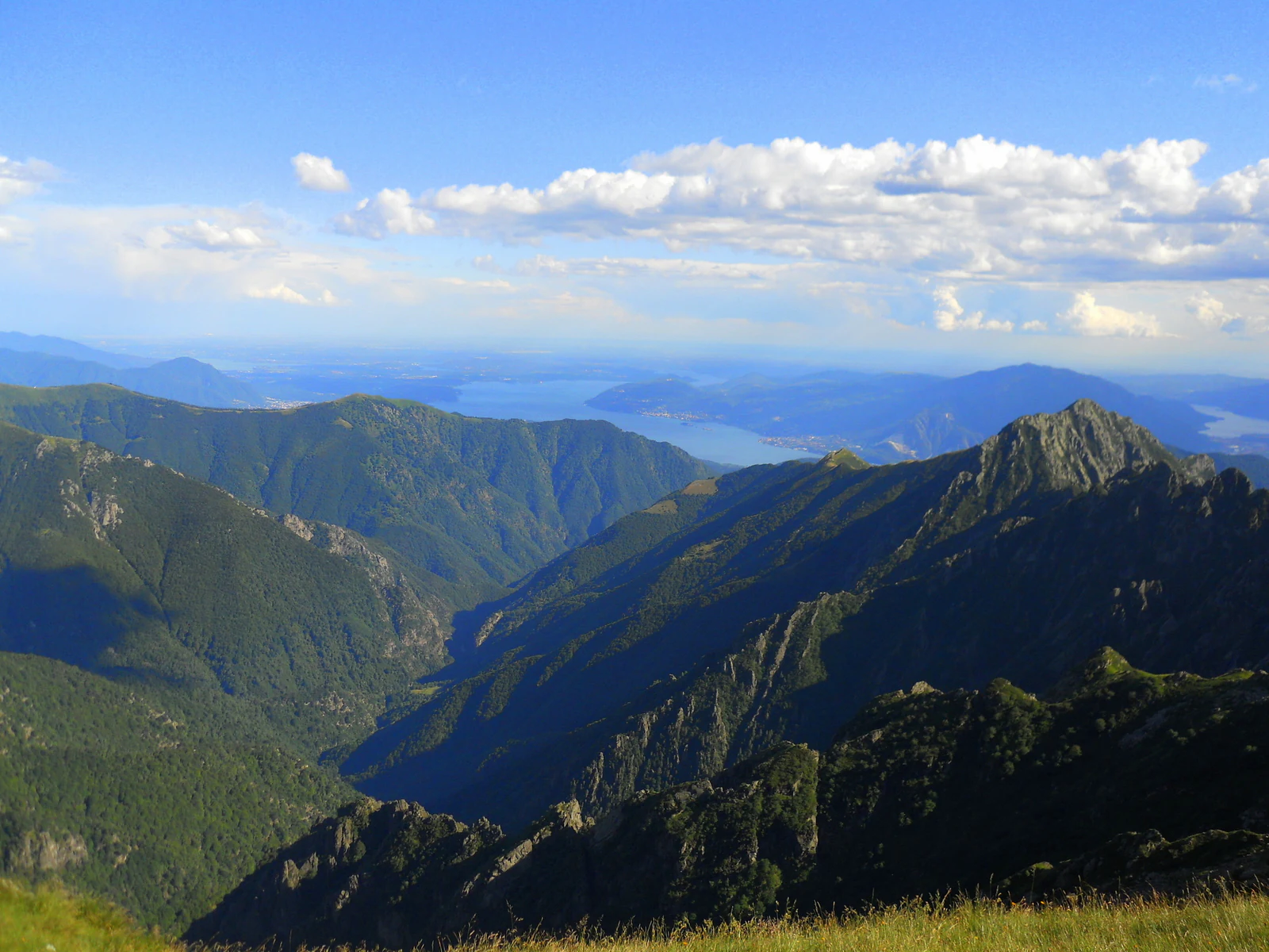 Panoramic landscape view of mountain ranges, valleys, and a distant lake under a blue sky with scattered clouds.