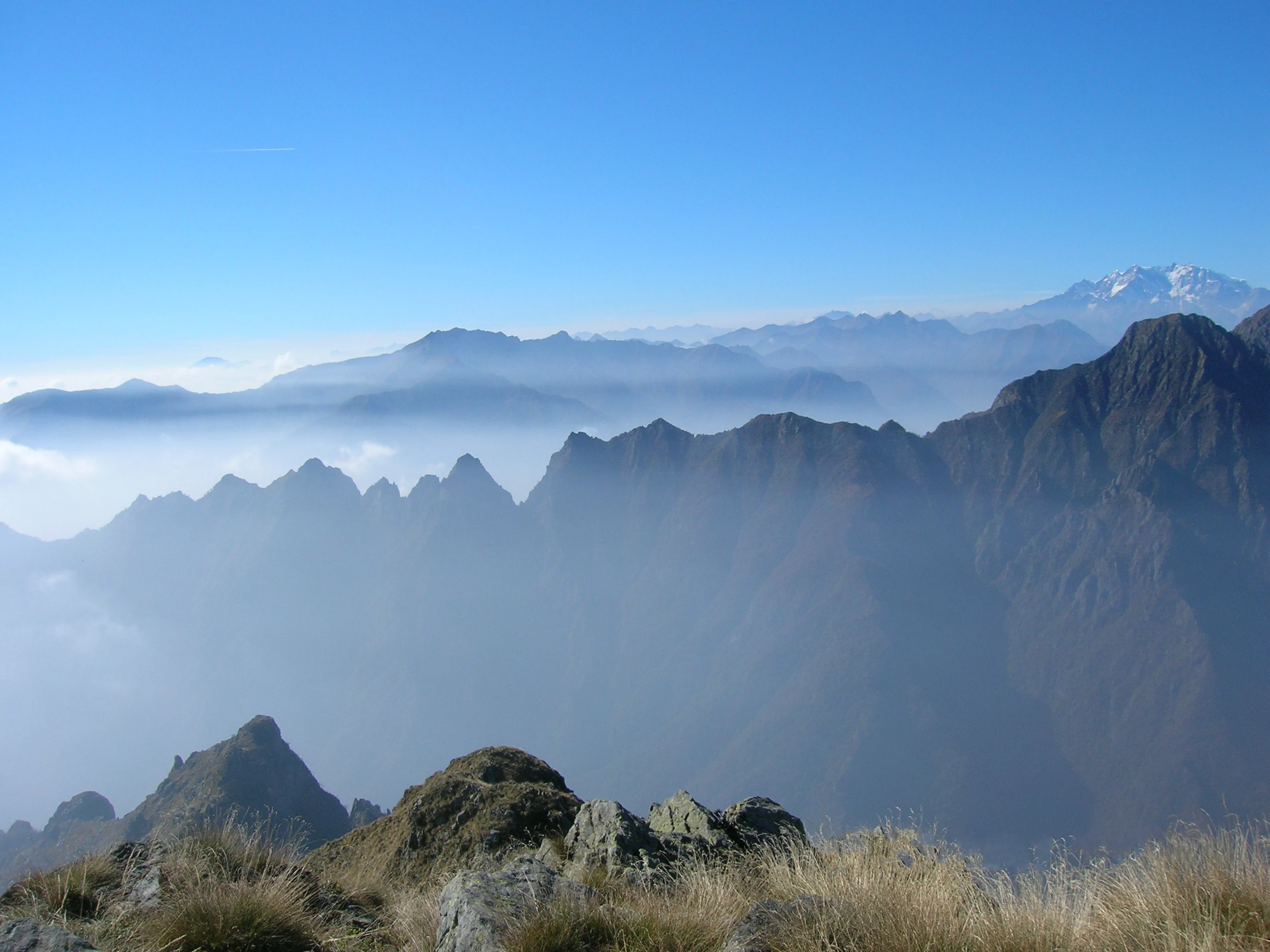 Mountain landscape with multiple peaks, misty valleys, and clear blue sky, viewed from a rocky hilltop with sparse vegetation