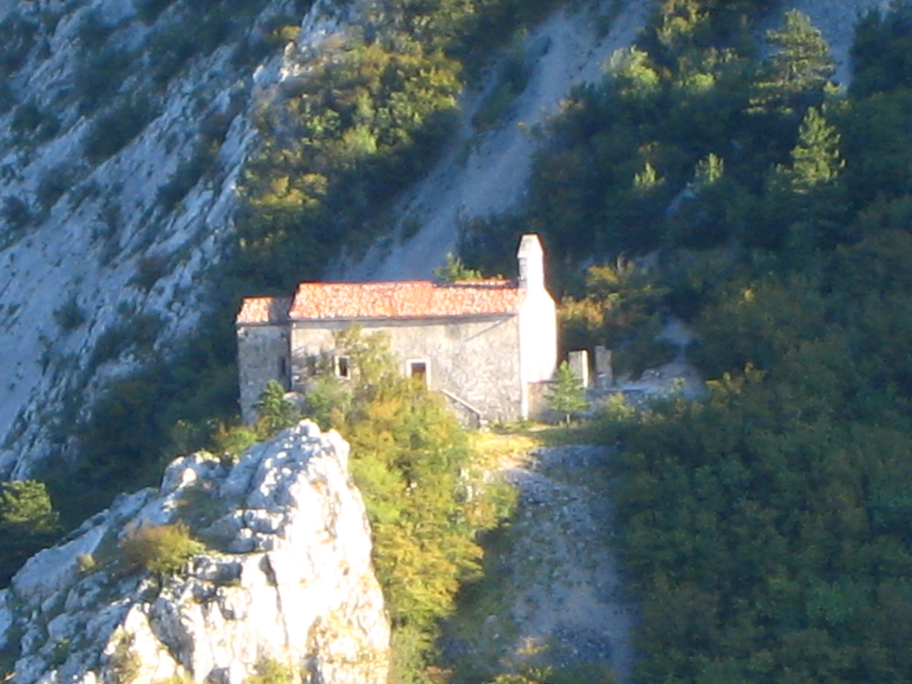 A small, historic stone church with a red-tiled roof sits on a hillside, nestled between steep limestone cliffs and dense vegetation in Val Rosandra.