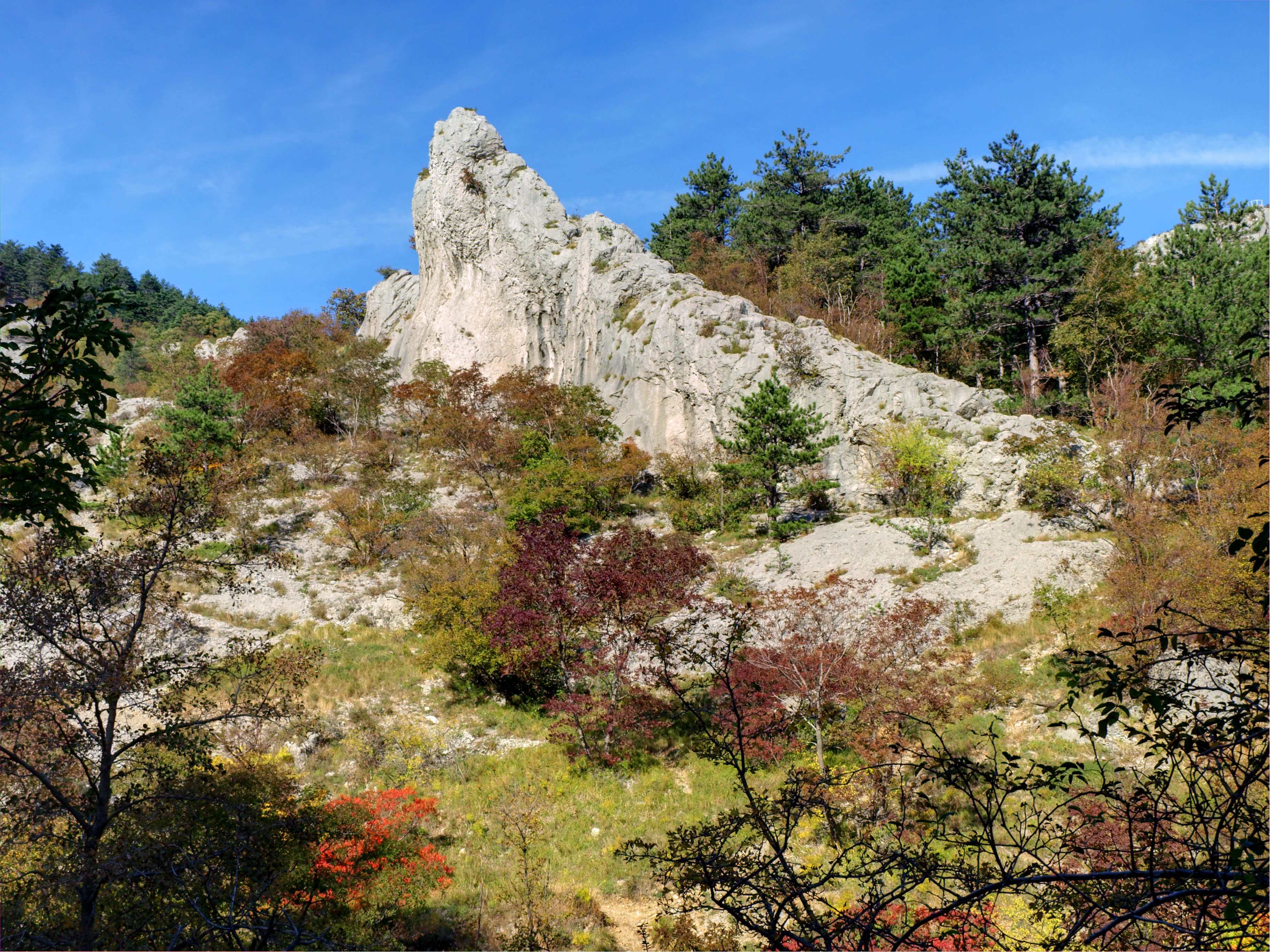 A jagged, light-colored limestone rock formation rises above a hillside covered in sparse trees and shrubs displaying autumn colors under a clear blue sky.
