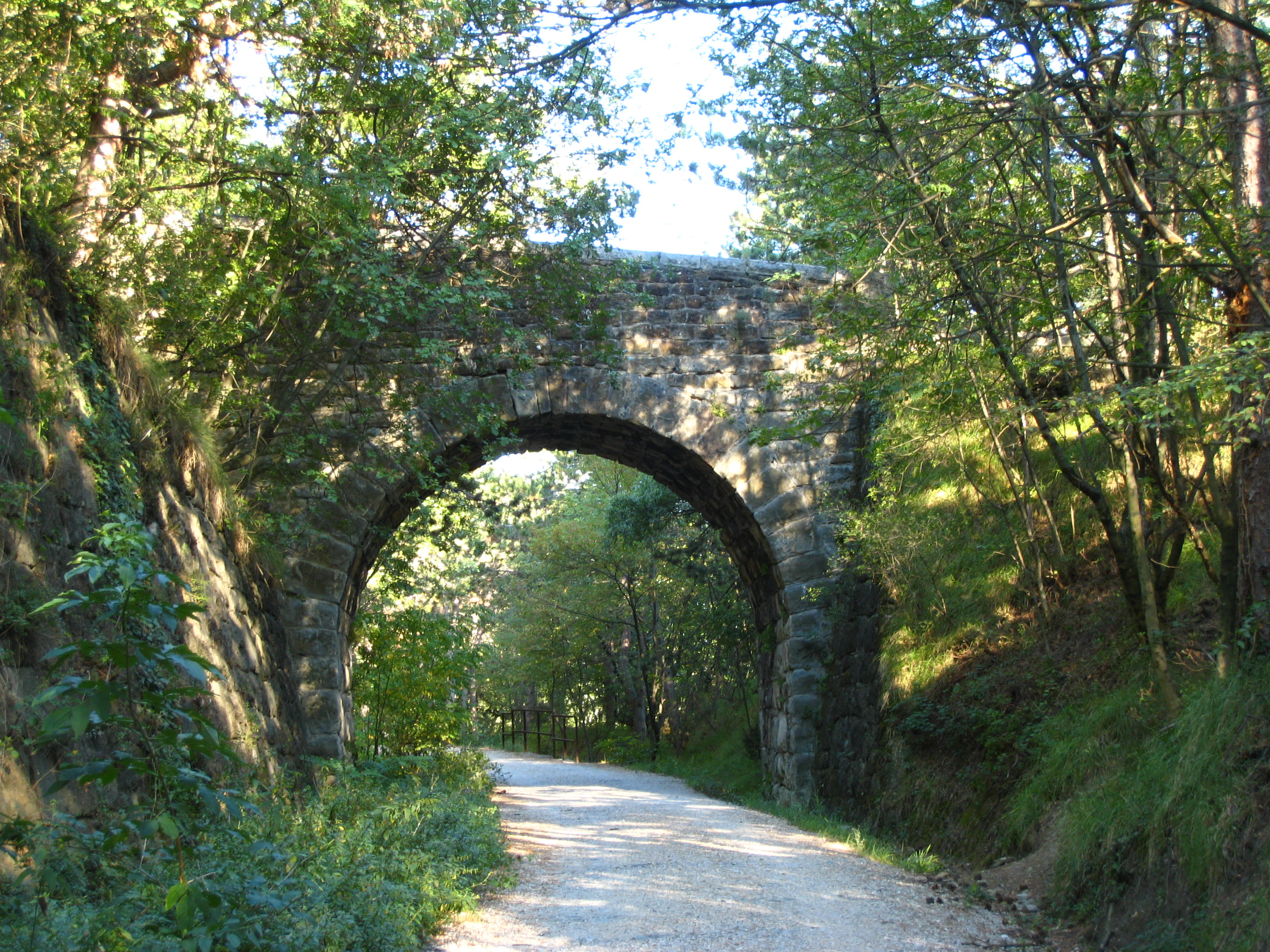 A gravel hiking trail runs through a wooded area under a historic stone arched bridge.