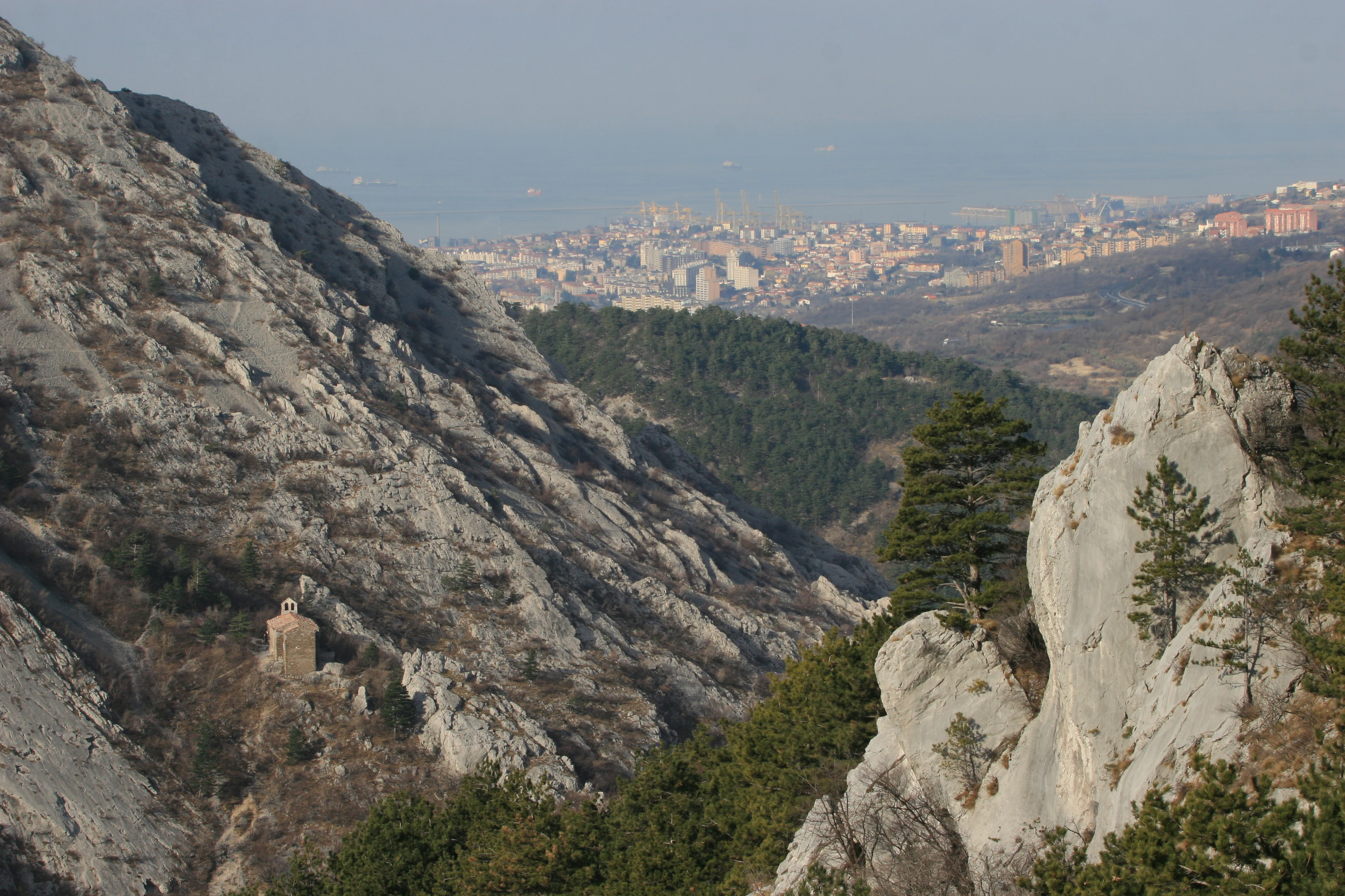 A wide angle view of a steep rocky valley with sparse trees in the foreground, looking out toward a distant urban cityscape under a hazy sky.