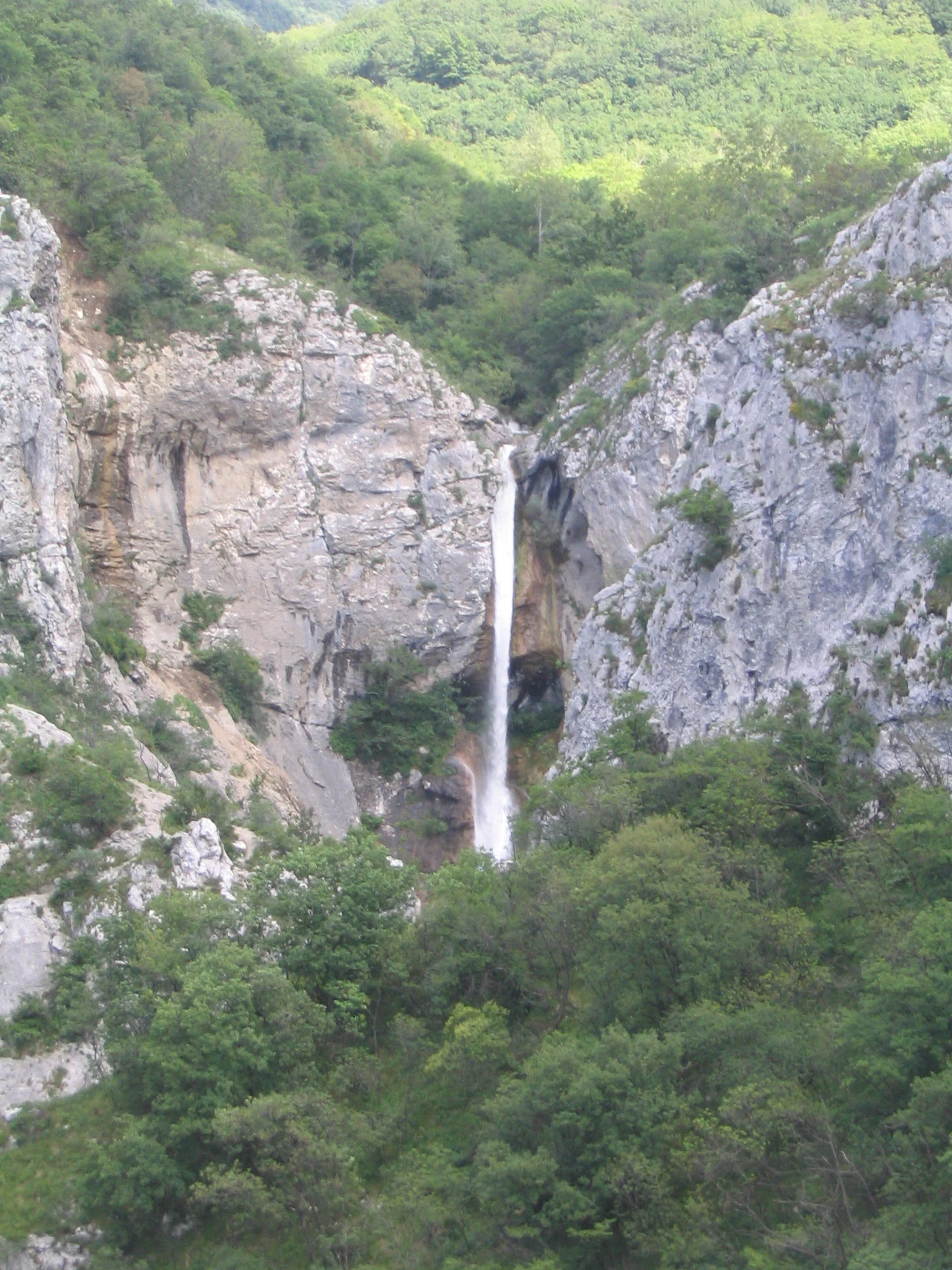 A waterfall flows down a vertical cliff face surrounded by dense green forest in a rocky valley.