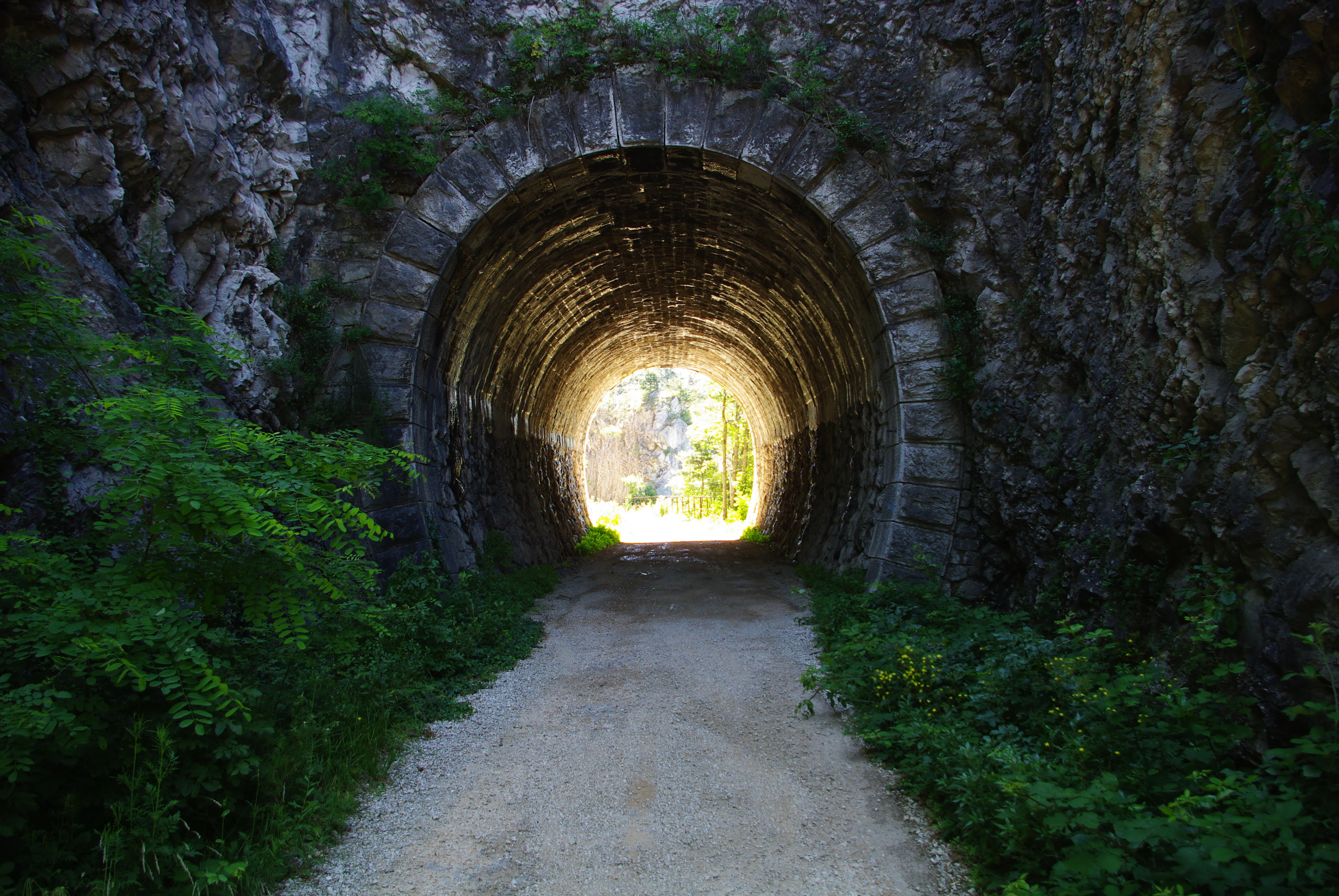 A view through a stone tunnel with a natural stone archway, looking toward a bright, sunlit exit framed by lush green foliage.
