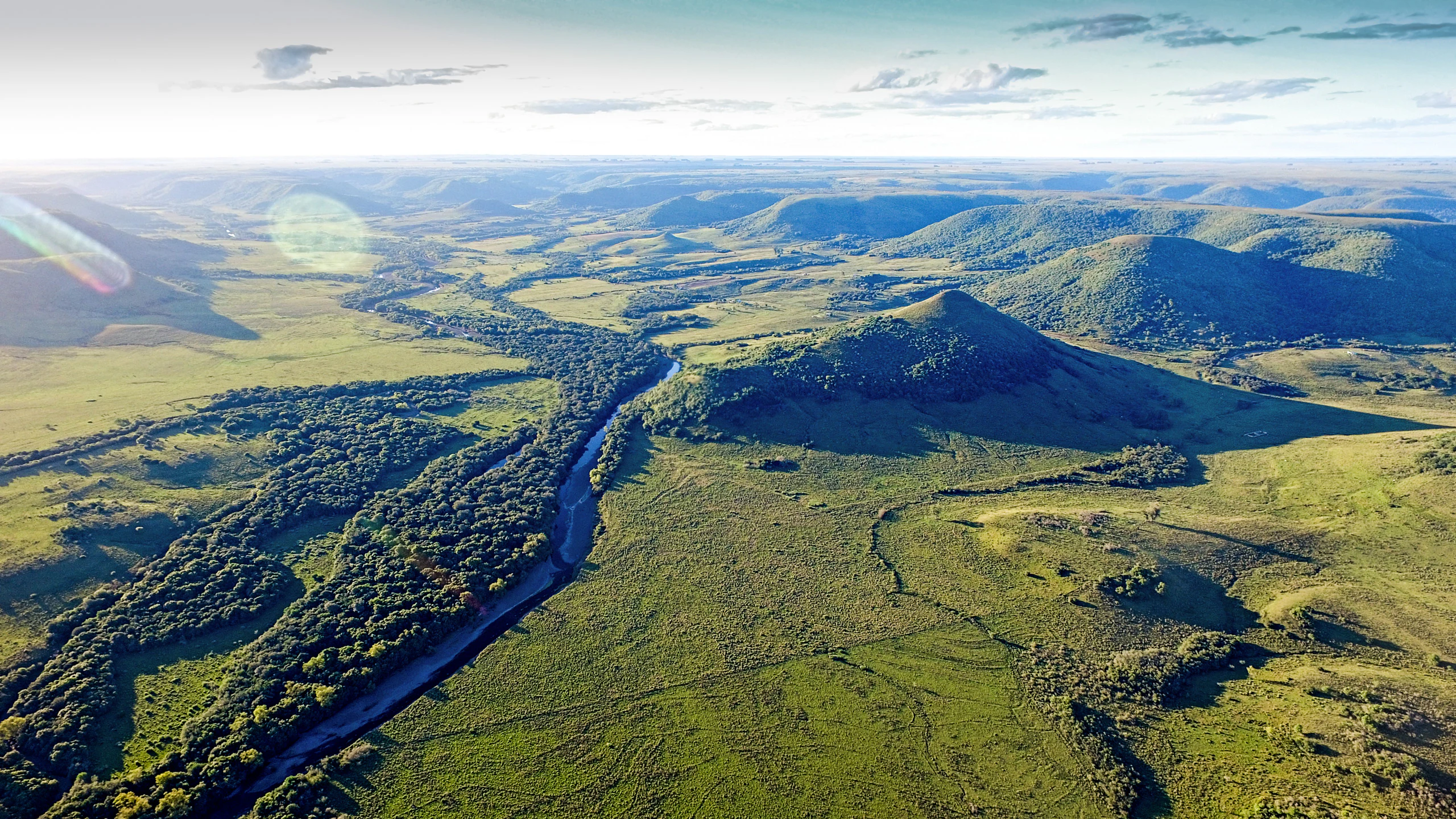 Vista panorámica aérea de un valle verde y exuberante en el norte de Uruguay, con un río serpenteante, corredores boscosos y colinas basálticas circundantes bajo un cielo brillante.