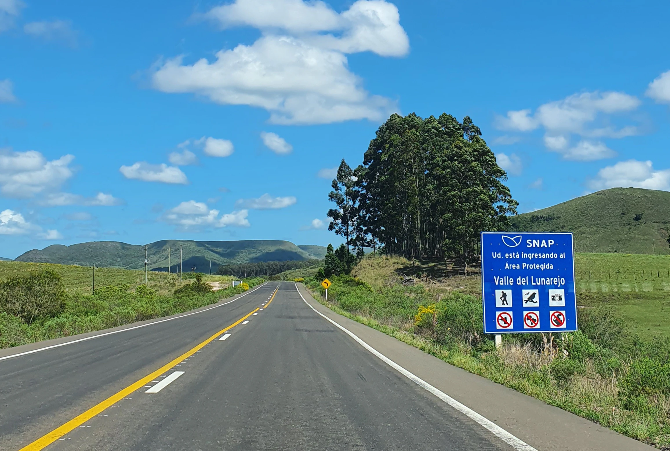 Una carretera asfaltada que conduce a un valle rural bajo un cielo azul con nubes dispersas. Una gran señal informativa azul al lado derecho indica la entrada al área protegida del Valle del Lunarejo.