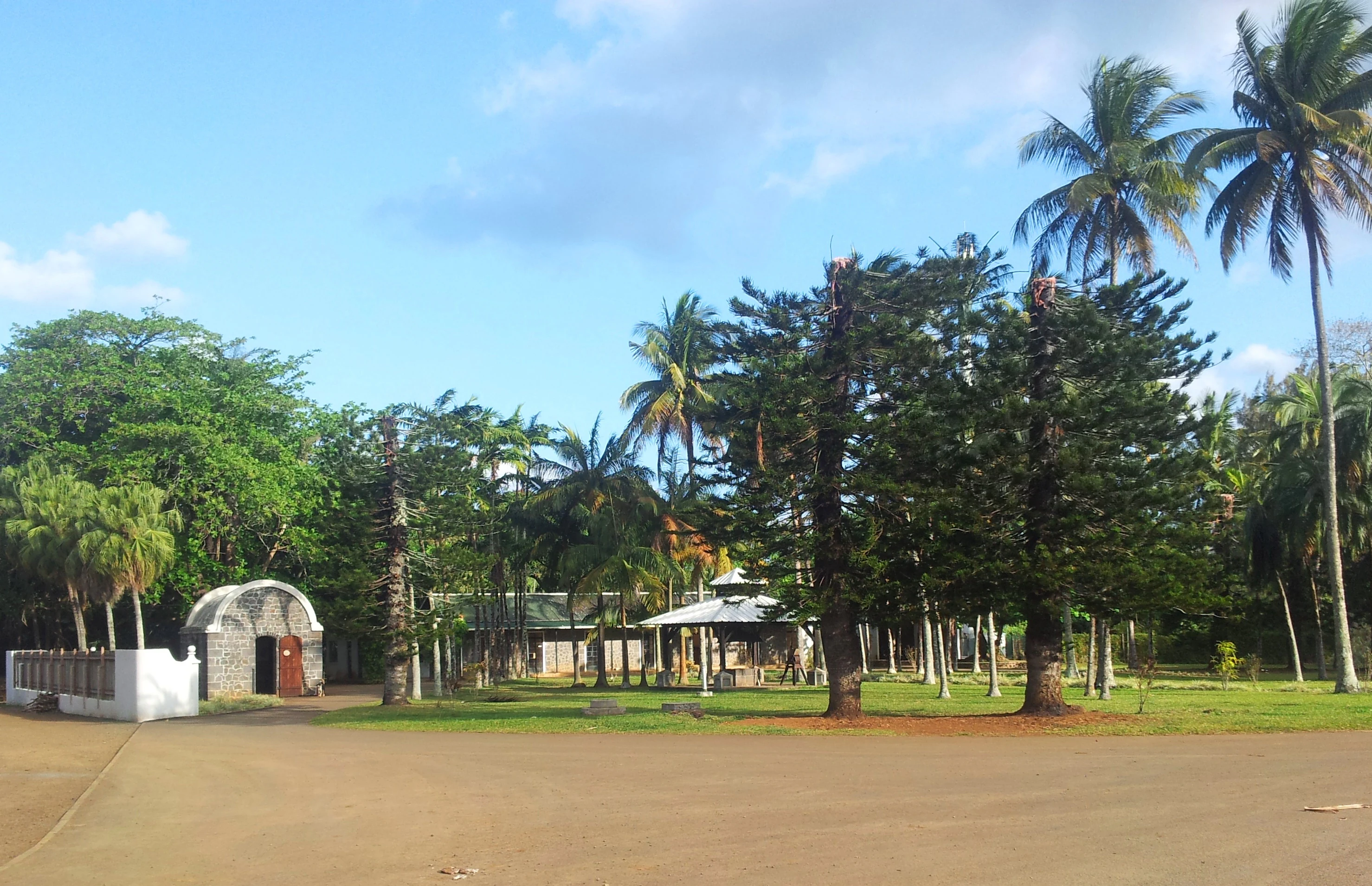 A view of historic stone and white-walled farm buildings surrounded by palm trees and pine-like trees in a garden setting.