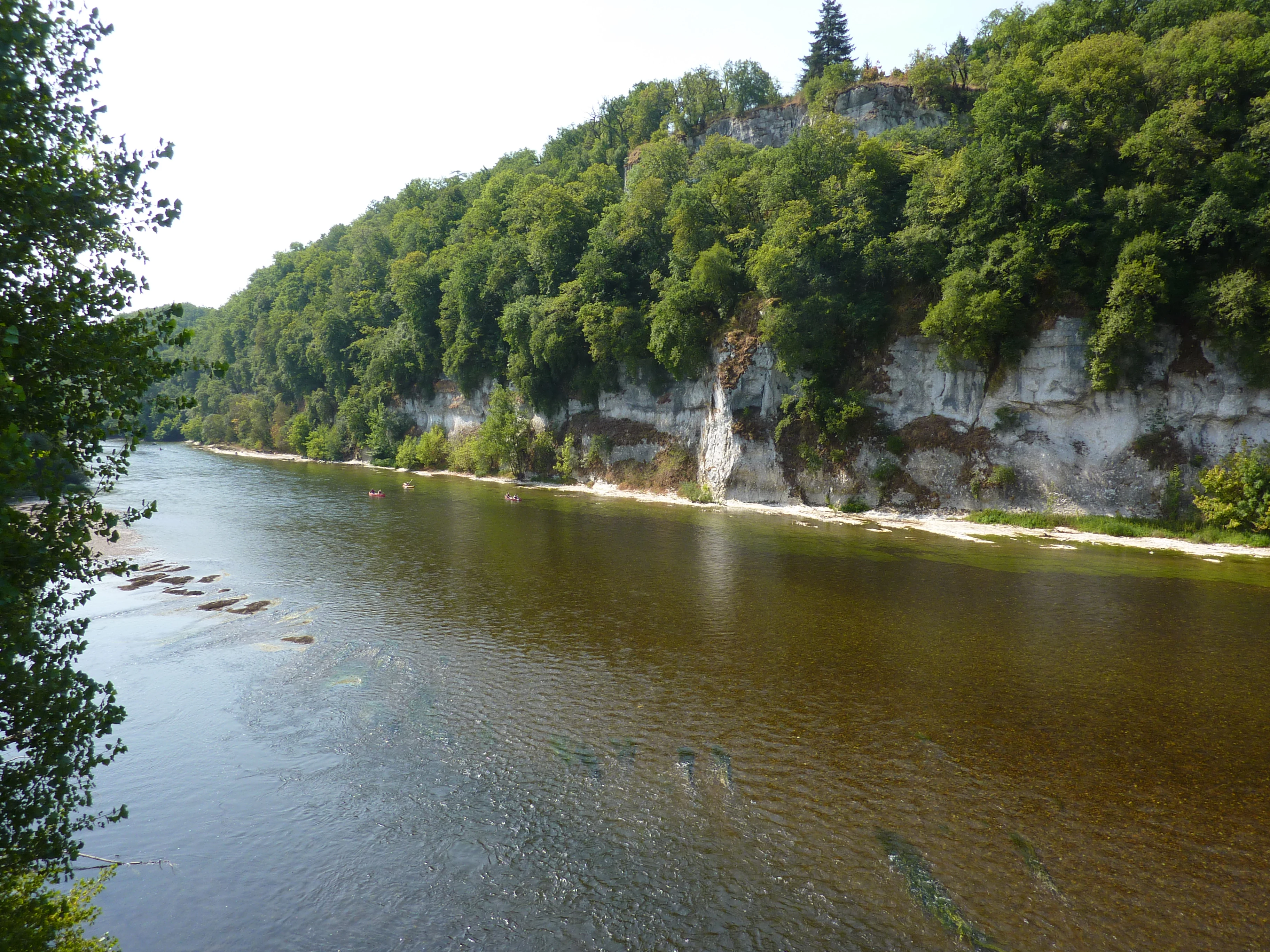 A wide river flows past a tall, forest-covered limestone cliff, with visible sandbars in shallow water during summer.