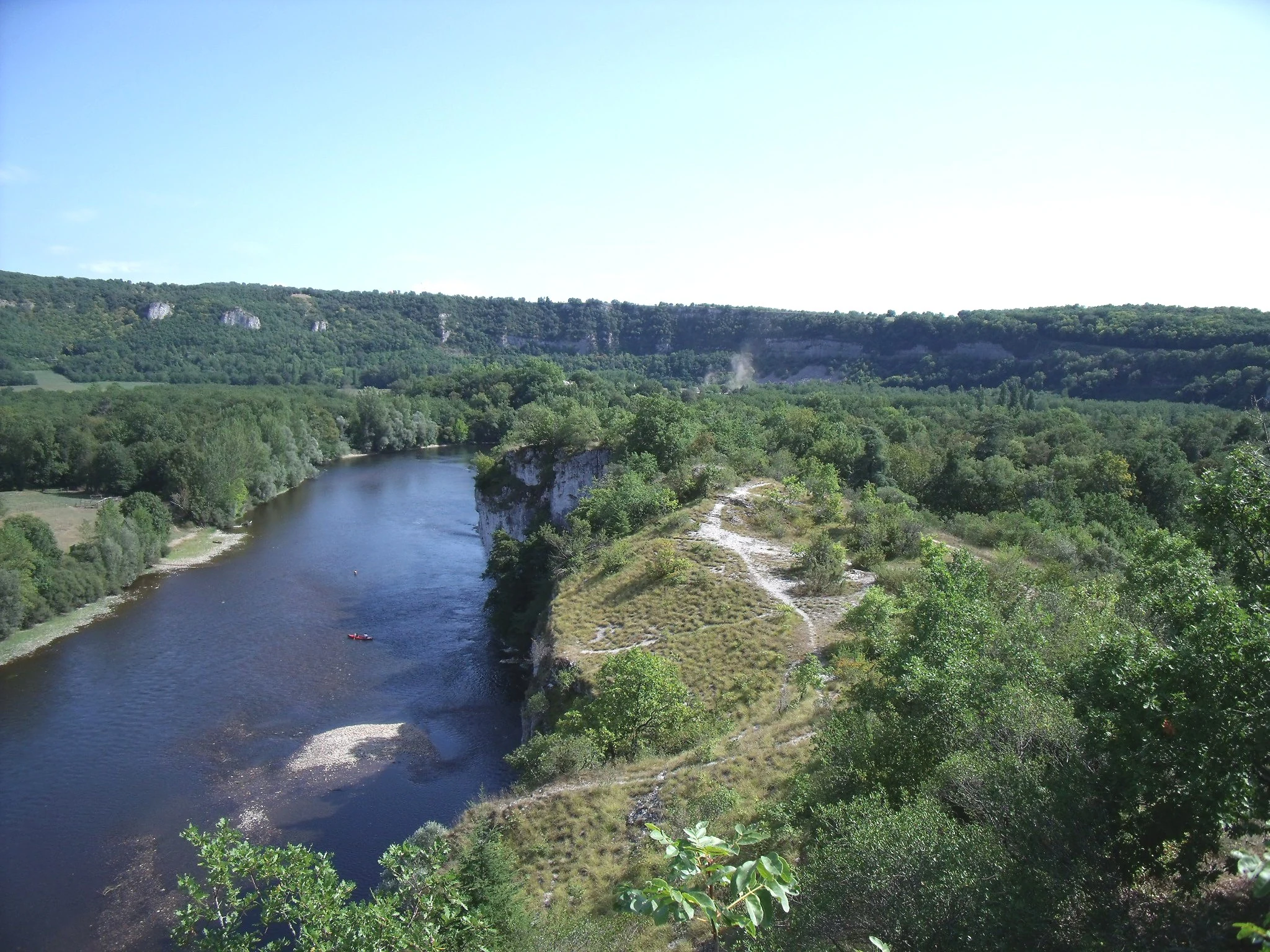 A wide, high-angle landscape shot looking down at the dark blue Dordogne River flowing through a lush green valley with limestone cliffs in the background.