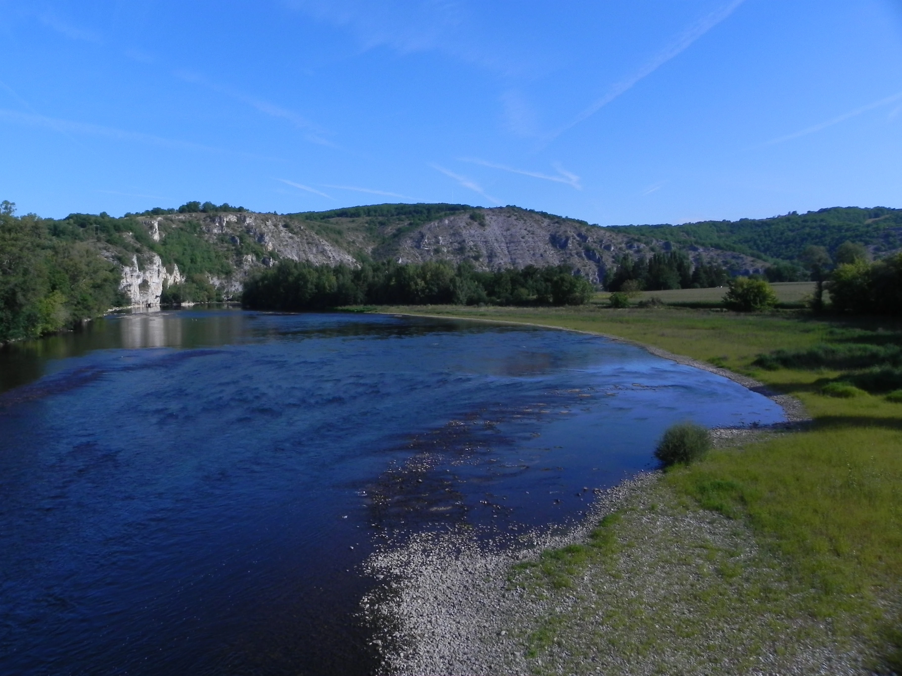 A wide, calm river bends through a valley lined with dense green forests and prominent light-colored limestone cliffs under a clear blue sky.