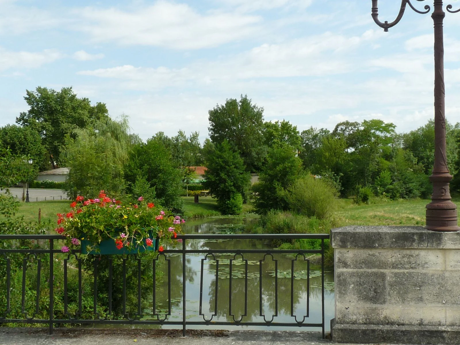 A view of the Tude River from a stone bridge featuring a decorative metal railing, a hanging flower basket, and a wrought-iron lamp post in the foreground.