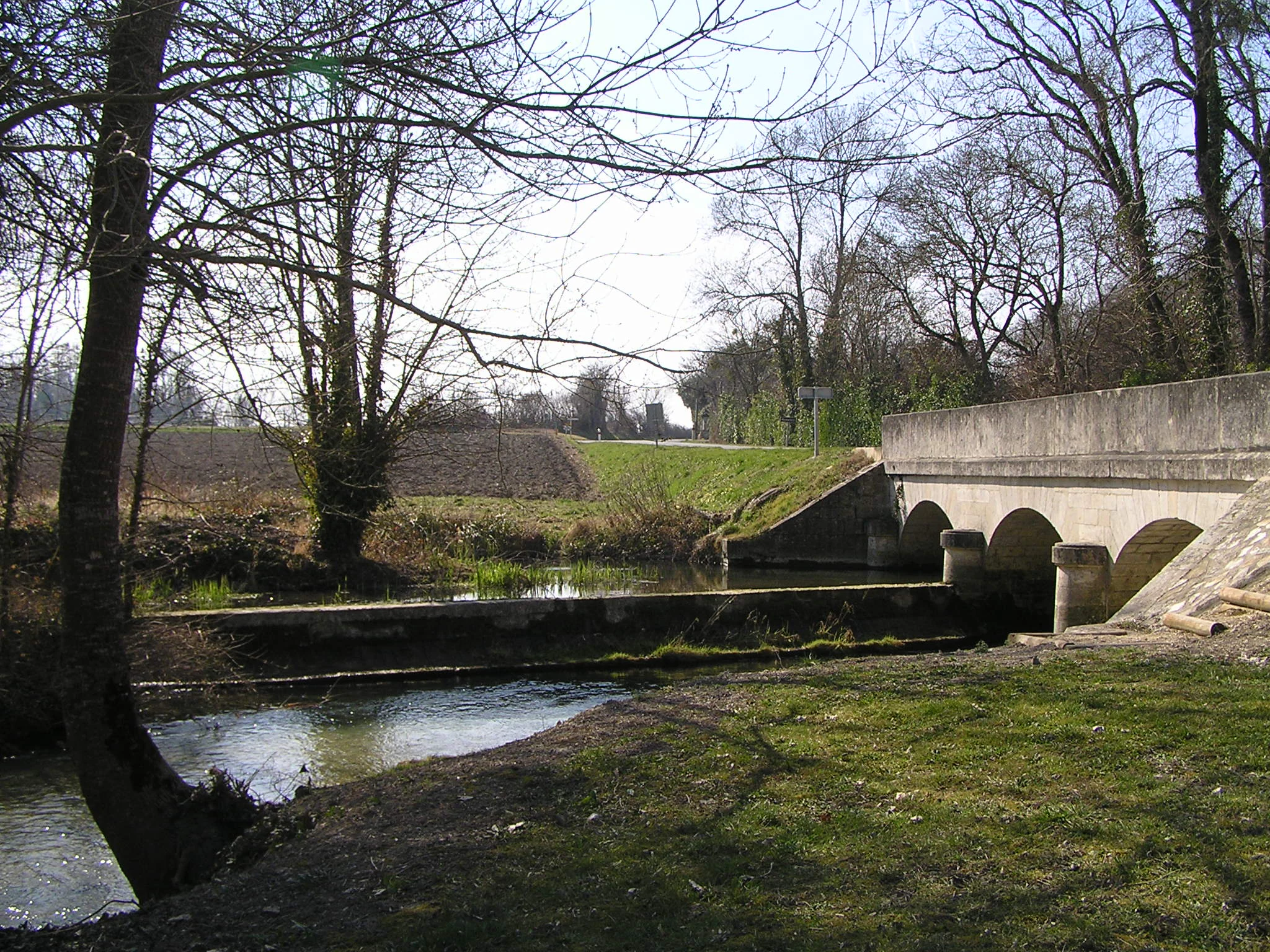 A small stone arched bridge crossing a calm river in a rural, wooded setting during a sunny day.