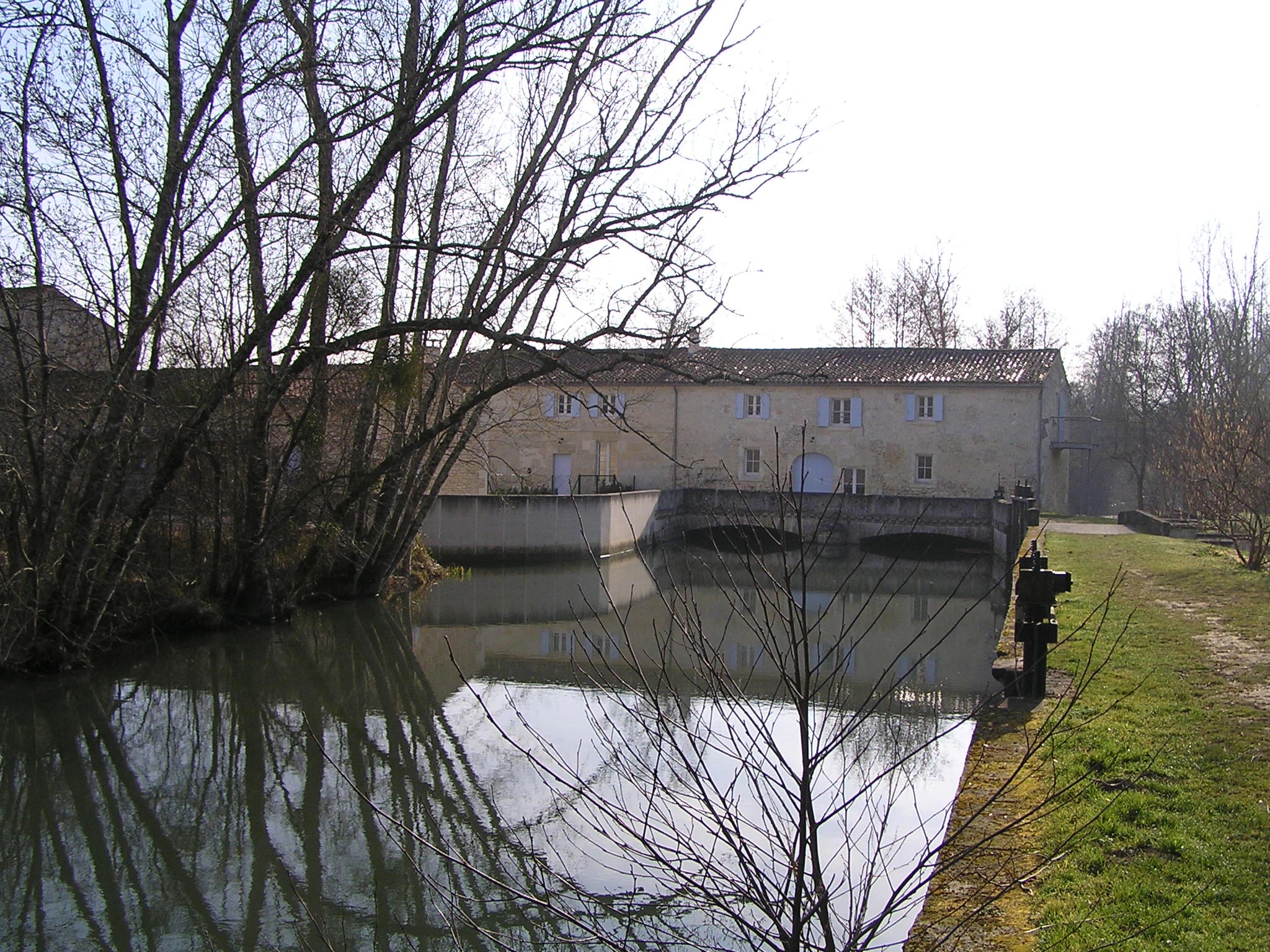 A calm section of the Antenne river in front of a stone building, identified as the Preziers mill, with a weir visible across the water and bare trees on the riverbank.