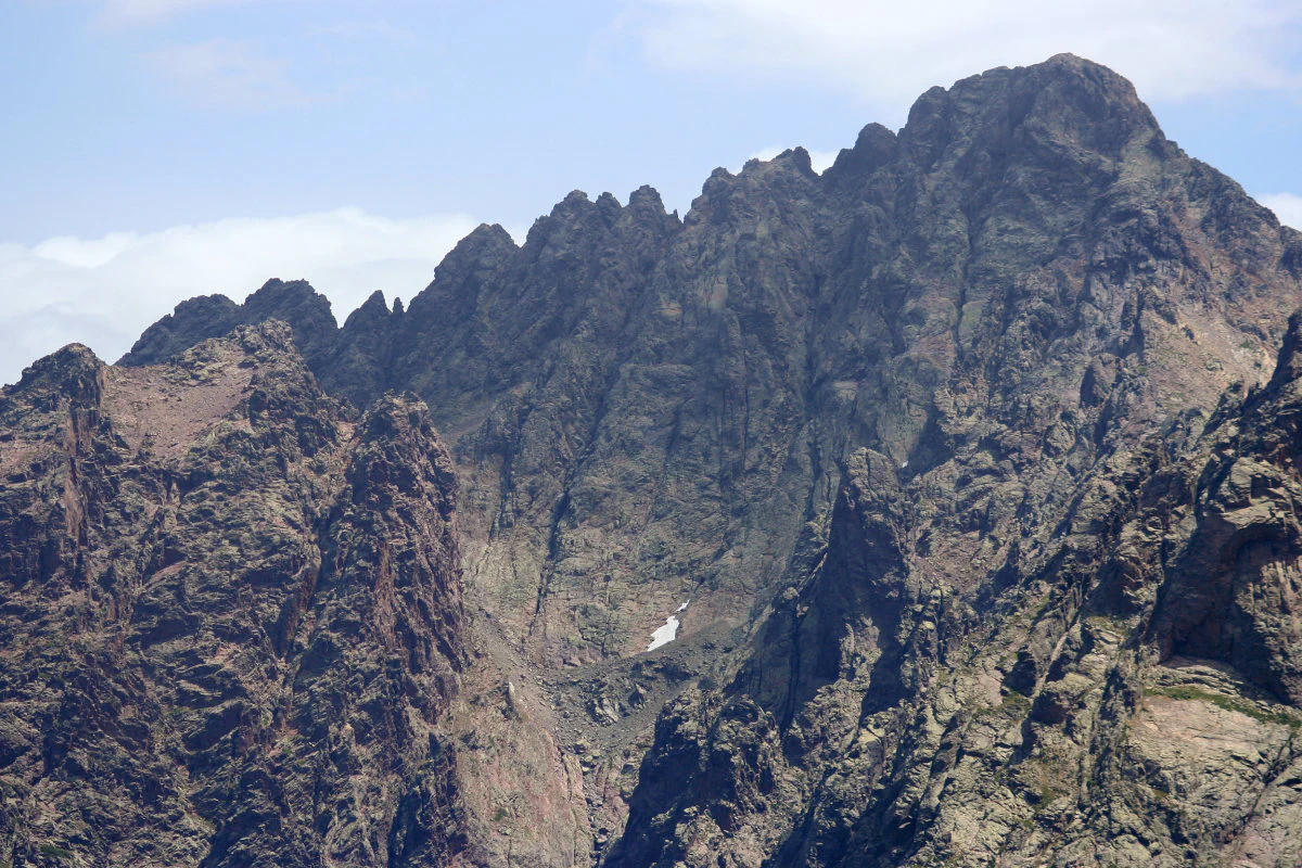 A jagged mountain peak with steep, dark rocky ridges and small patches of snow in the crevices under a partly cloudy blue sky.
