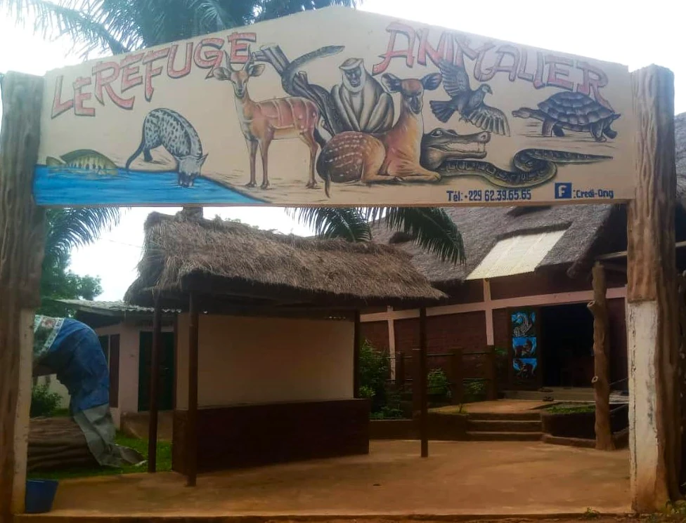 A large hand-painted wooden sign reading Le Refuge Animalier is suspended between two tree-trunk pillars, featuring colorful illustrations of various animals including a sitatunga, a primate, a crocodile, a snake, a bird, and a tortoise.
