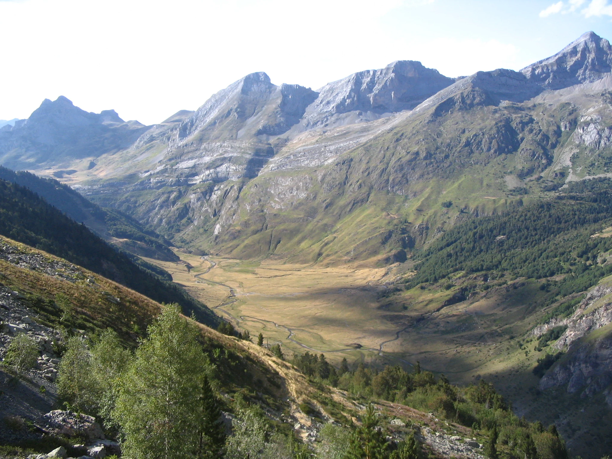 A high-angle, wide shot of a broad mountain valley in the Pyrenees featuring a winding stream through a grassy basin, surrounded by steep, rocky slopes and alpine ridges.