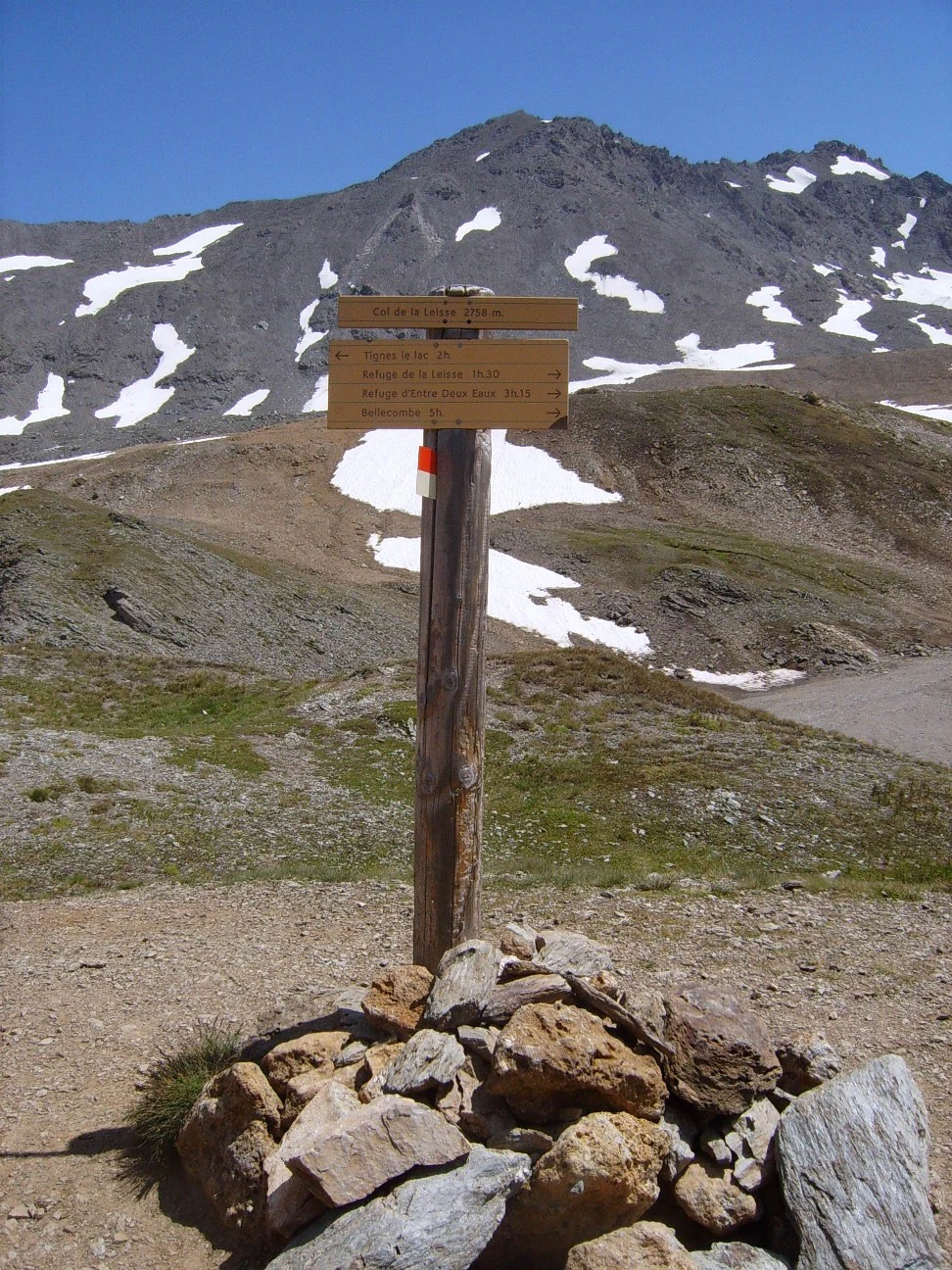 Wooden signpost with directional information at a mountain pass, surrounded by rocks, with snow patches on the mountains in the background