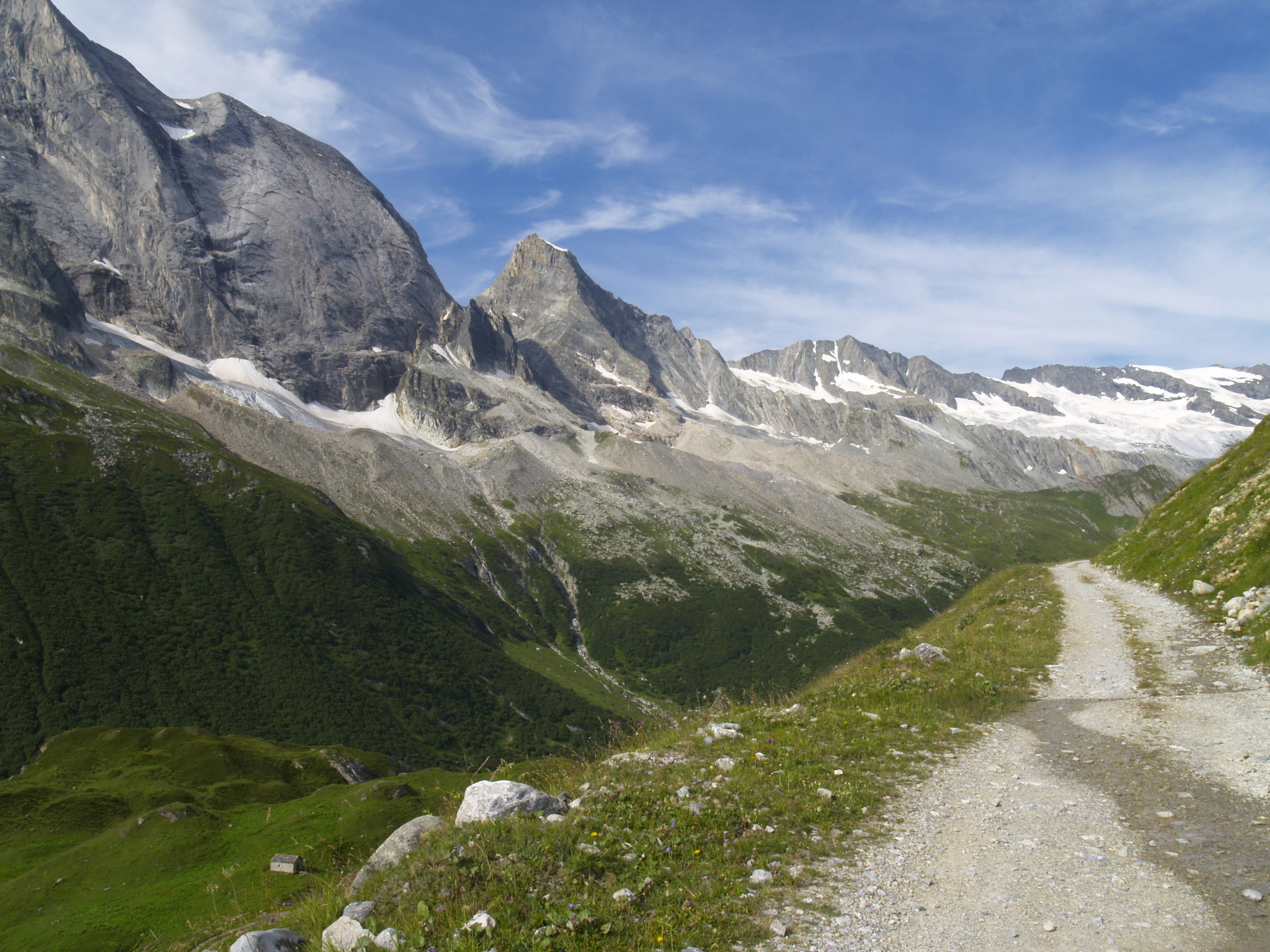 A gravel trail leads through a green valley with rocky mountains and glaciers in the background under a blue sky with scattered clouds