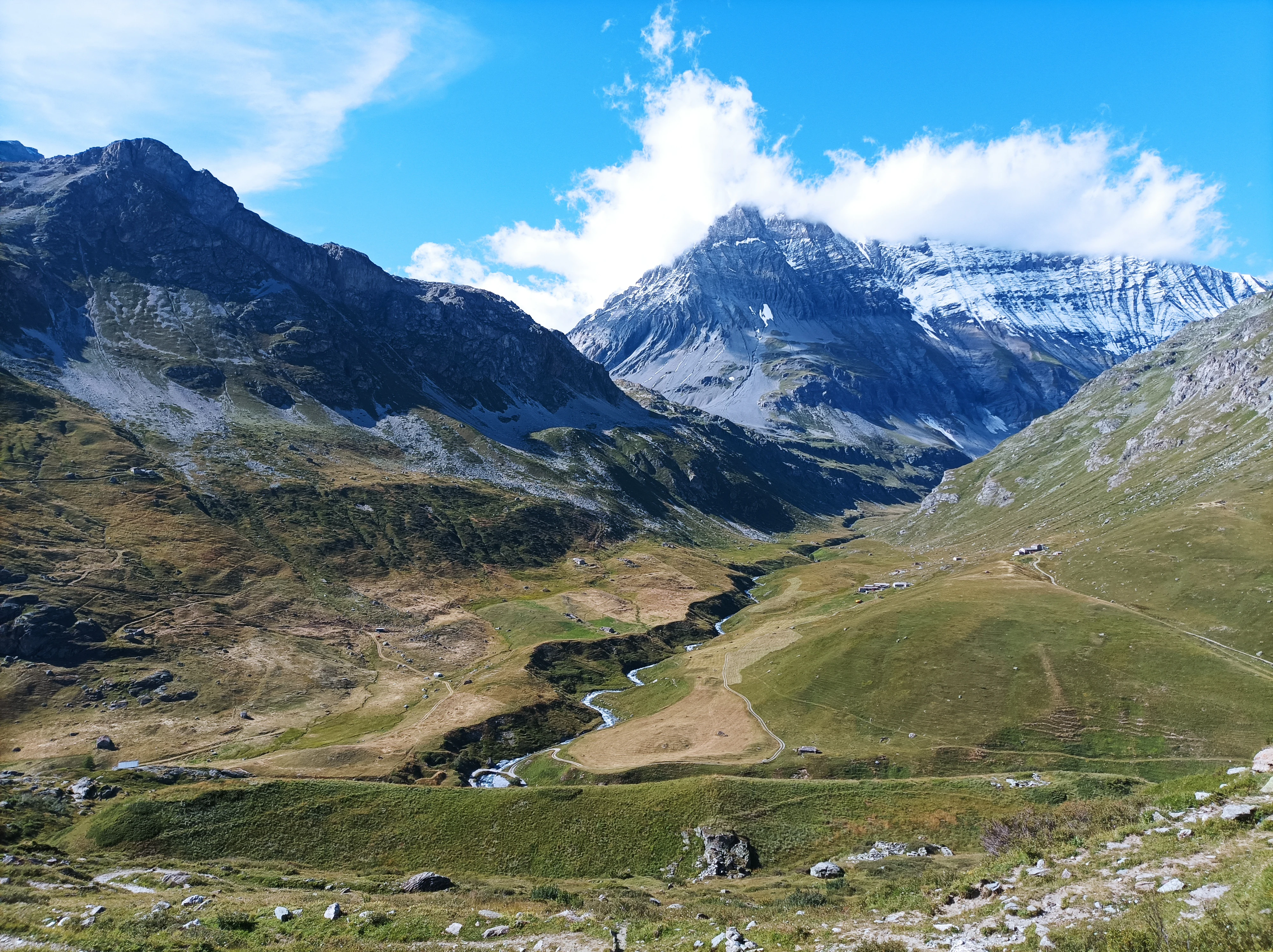 Wide landscape view of a mountain valley with Grande Casse peak in the background, green slopes, winding river, and scattered rocks under a blue sky with clouds.