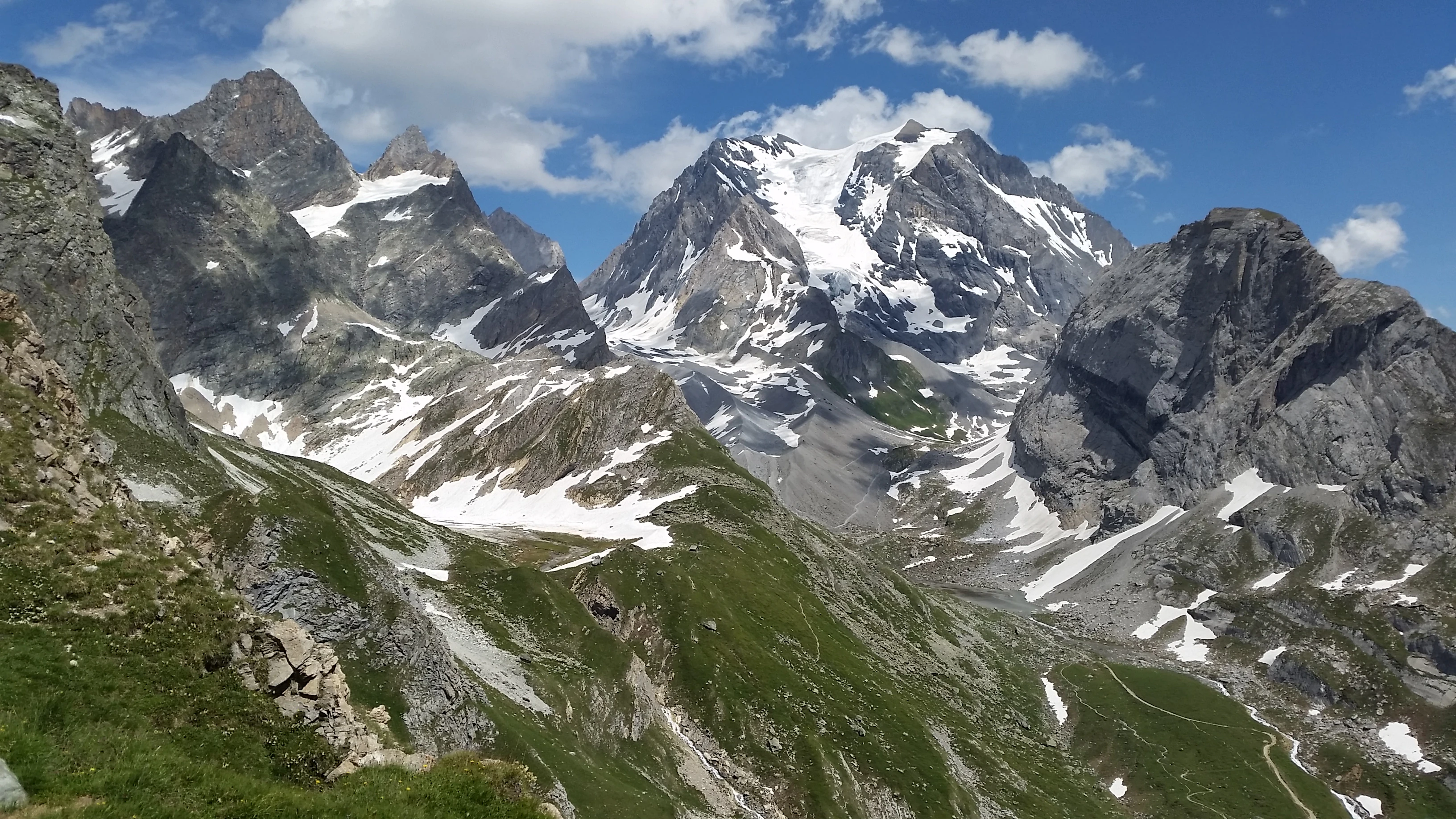 Snow-capped mountain peaks with green alpine meadows under a blue sky with scattered clouds
