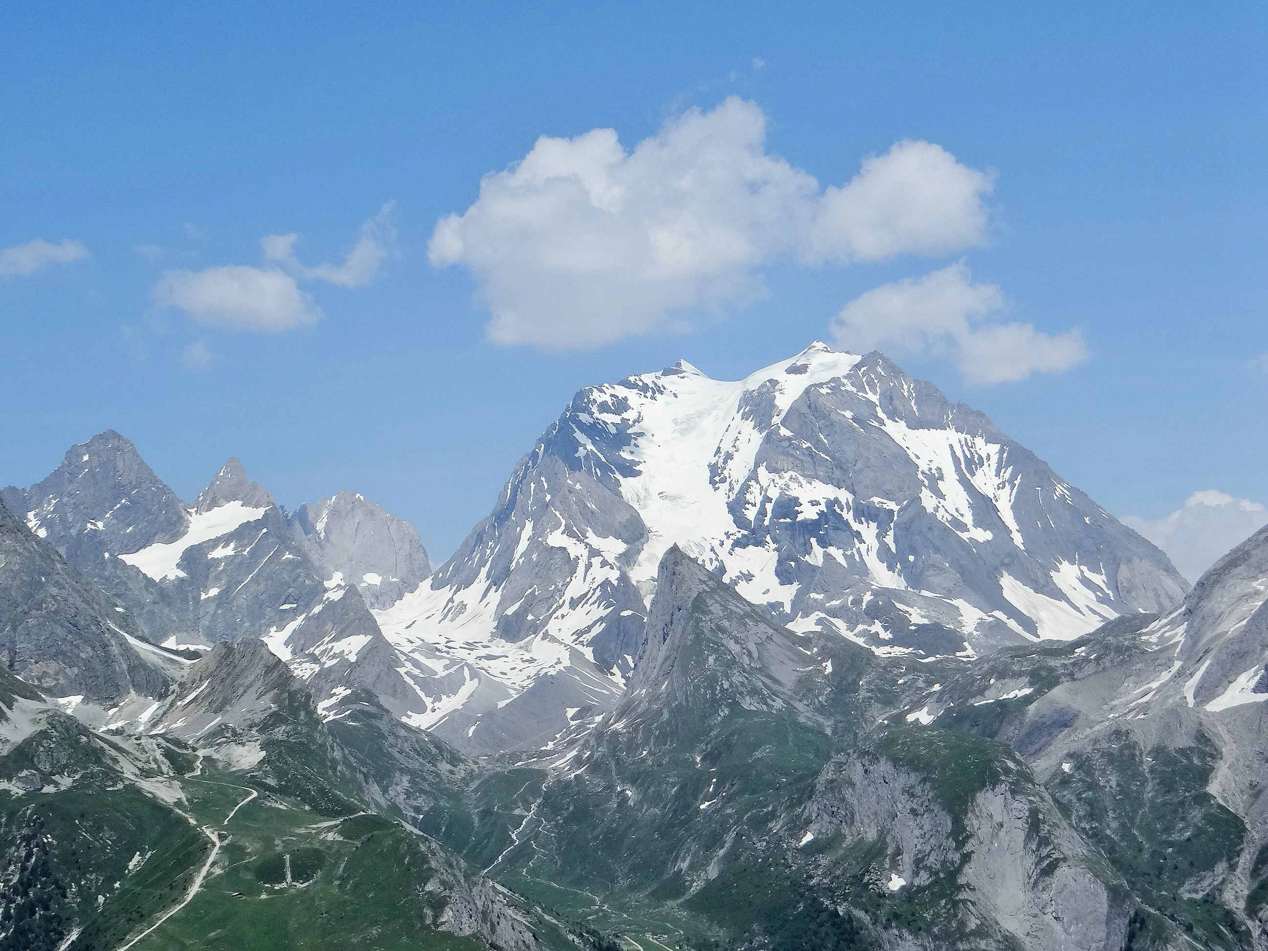 Snow-capped mountain peak with rocky slopes and green valleys under a blue sky with scattered clouds.